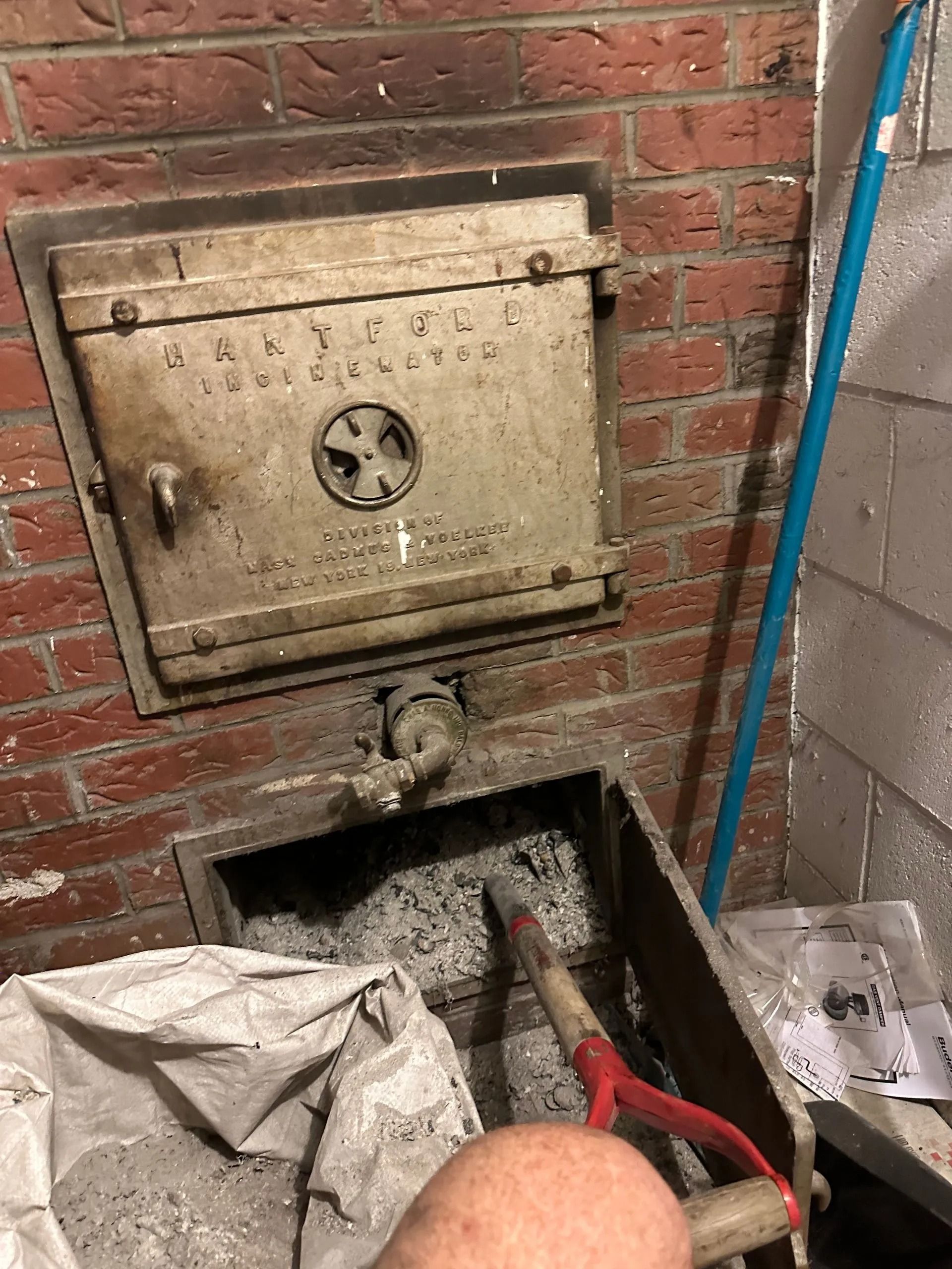 A metal furnace door in a brick wall, ash bin open, with ash, shovel, and blue broom visible.