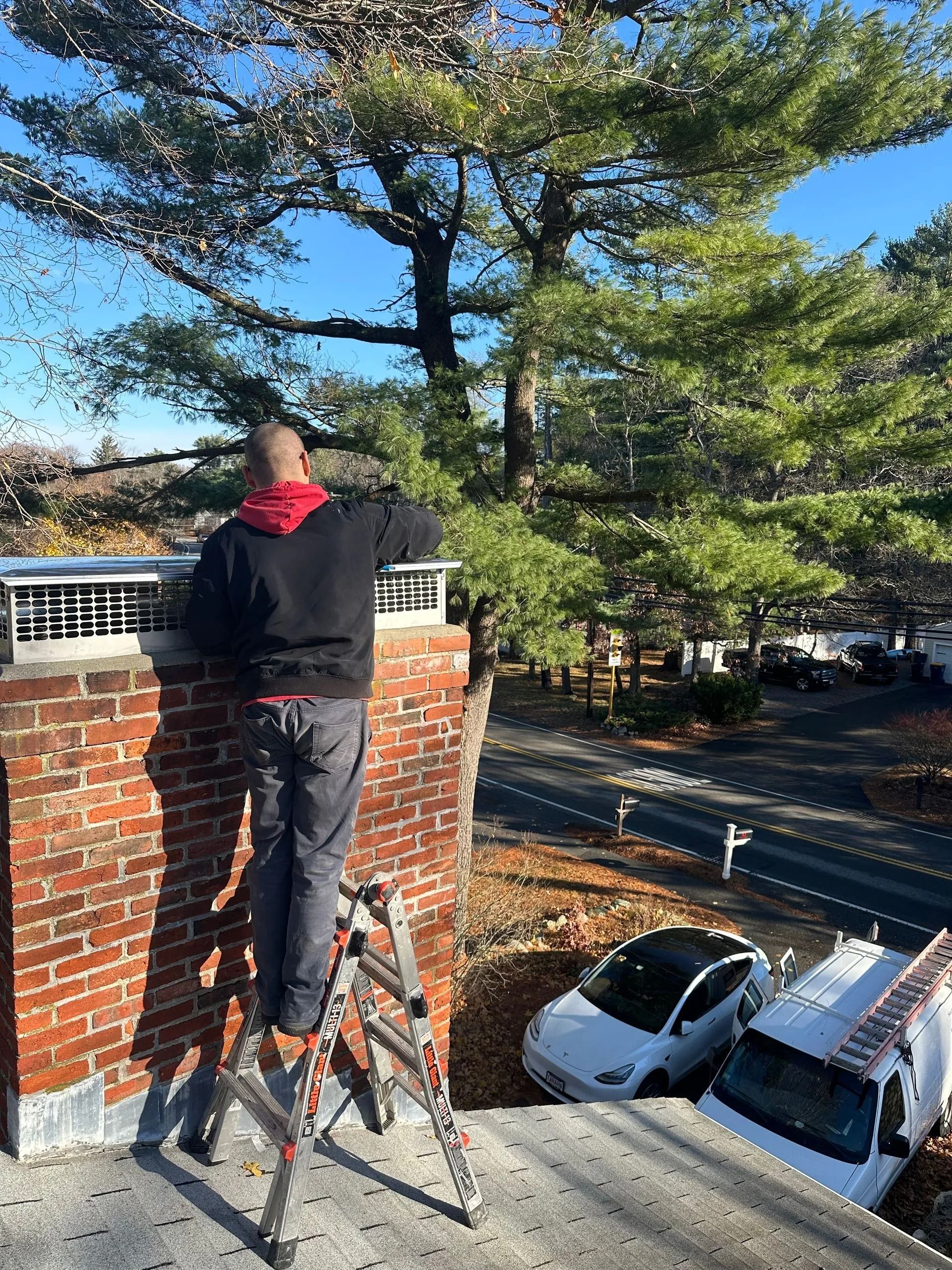 Person on a ladder, inspecting a brick chimney on a rooftop. A white Tesla and van parked below.