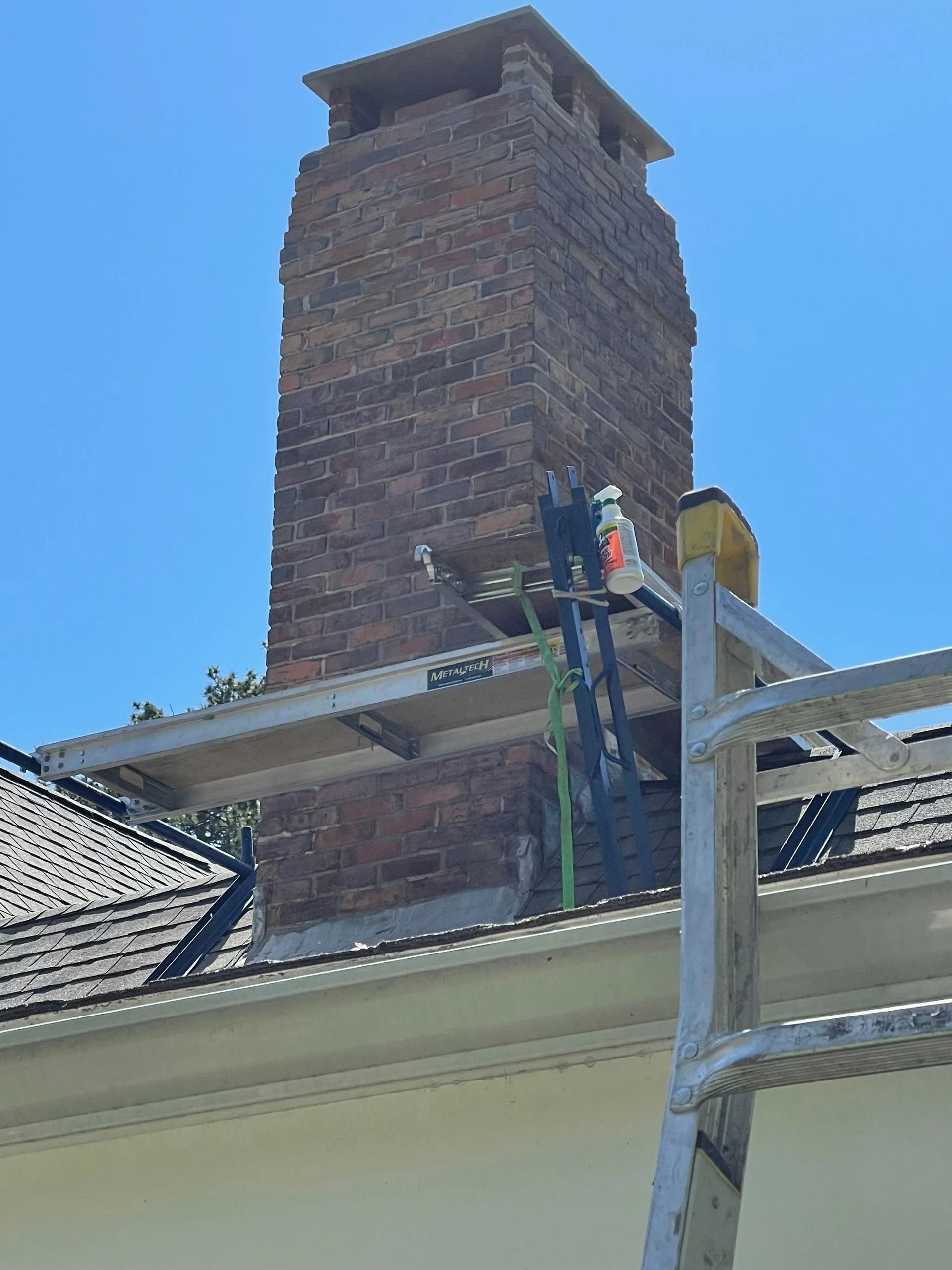 Brick chimney on a roof with scaffolding, being worked on under a blue sky.