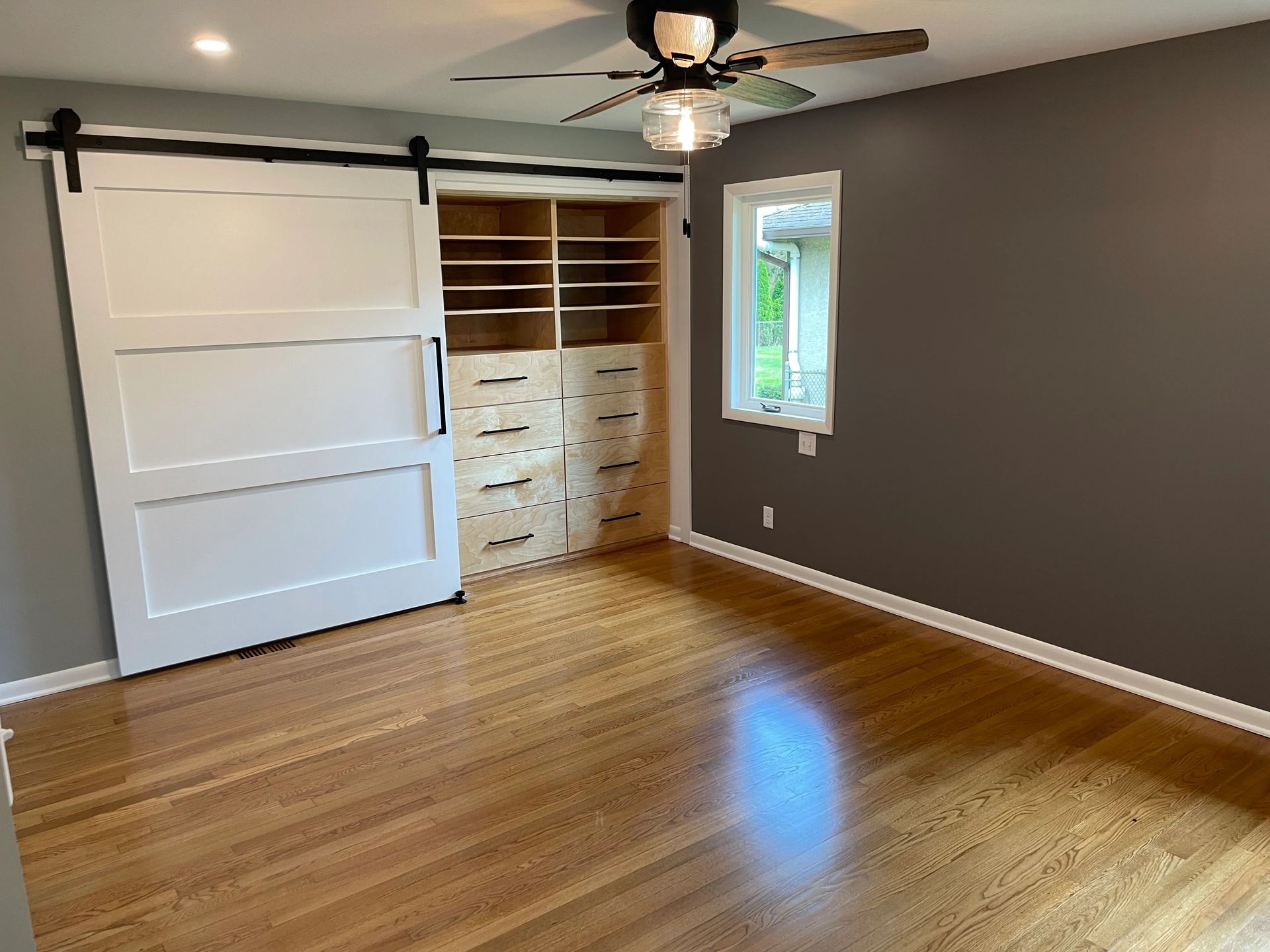 Bedroom with hardwood floors, a white sliding barn door, and gray walls. Window on the right.