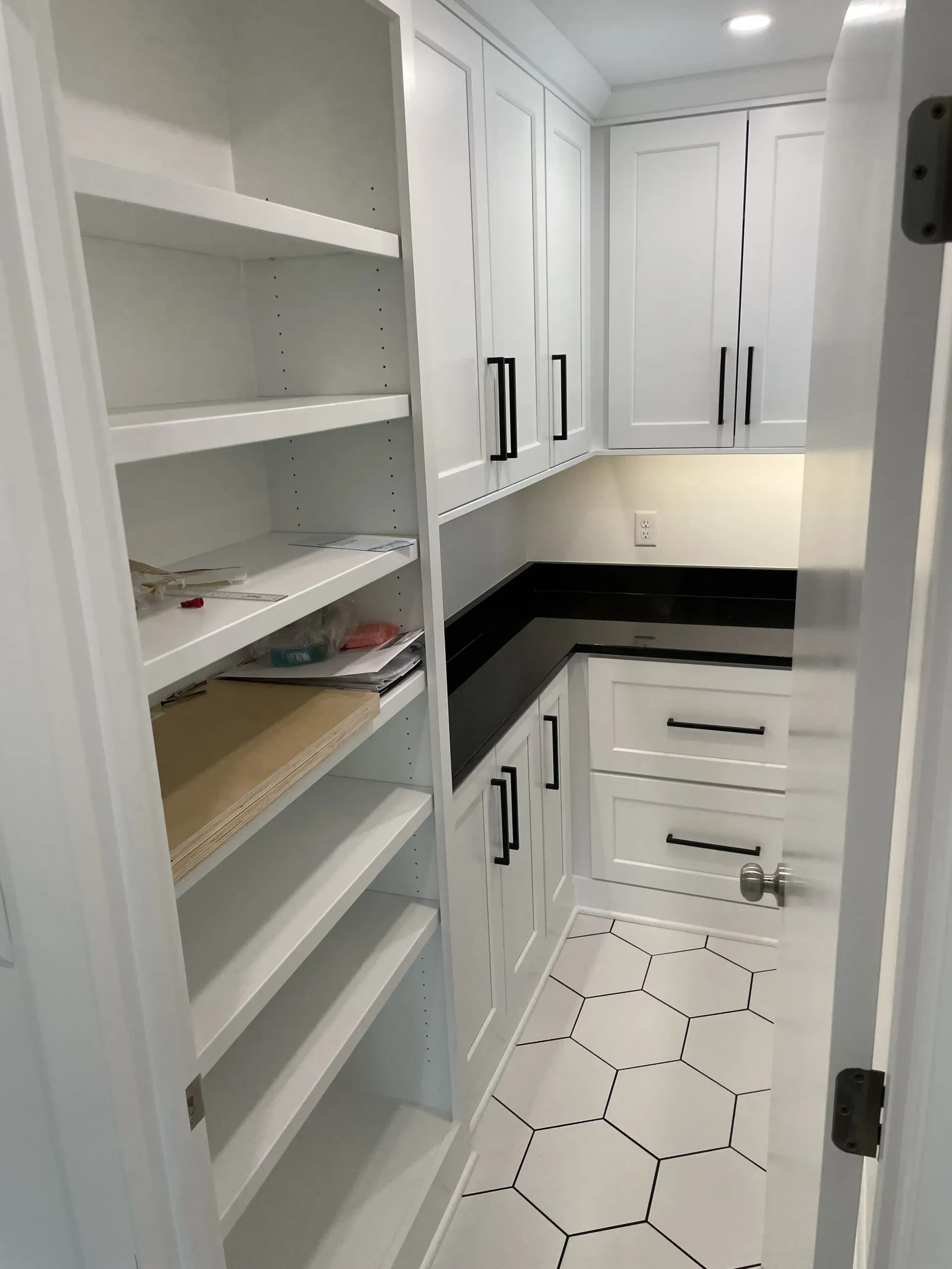 White pantry with shelves and cabinets. Black countertops and hardware. Hexagon tile floor.