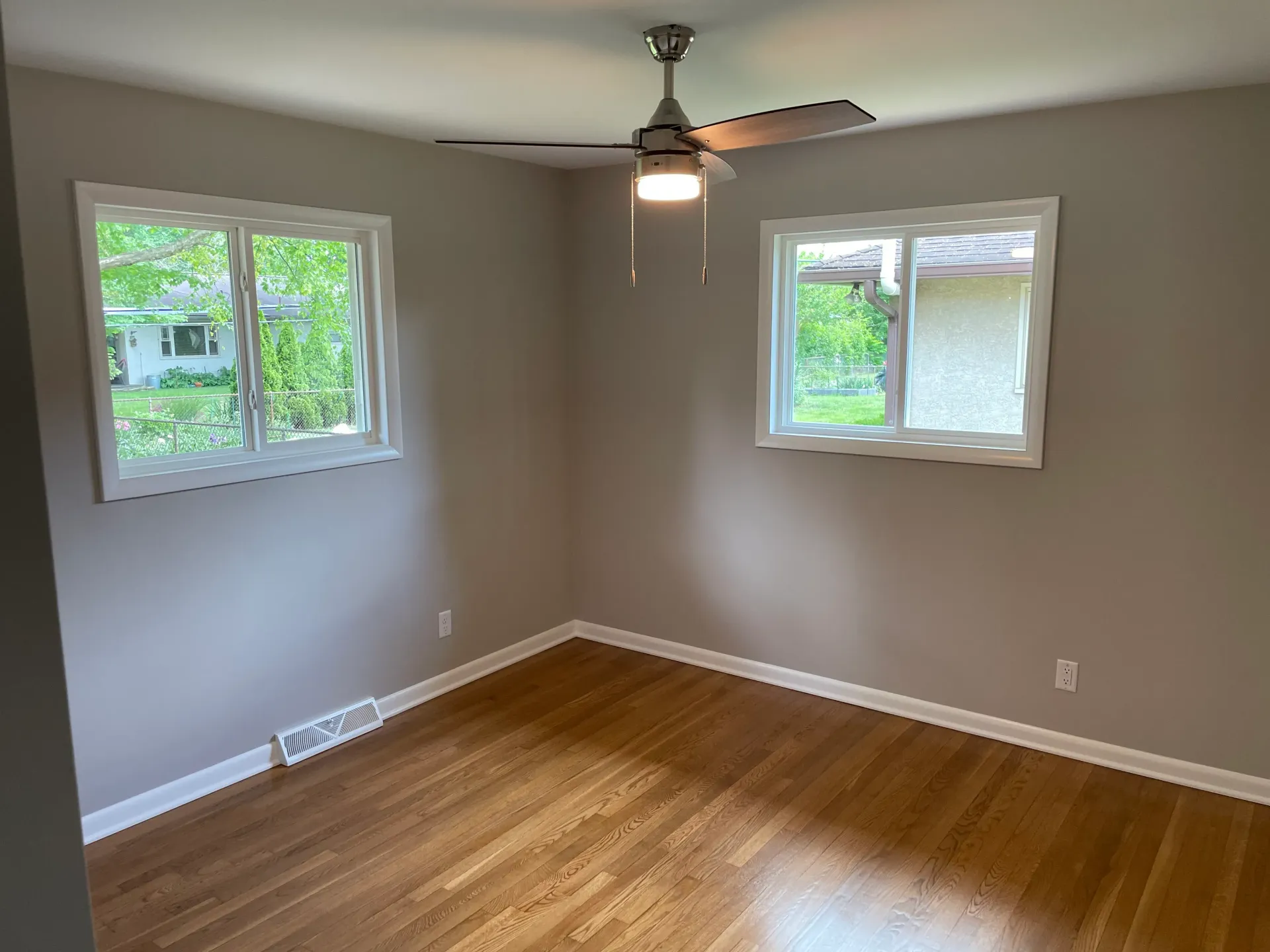 Empty bedroom with hardwood floors, two windows, and a ceiling fan. Walls are light grey.