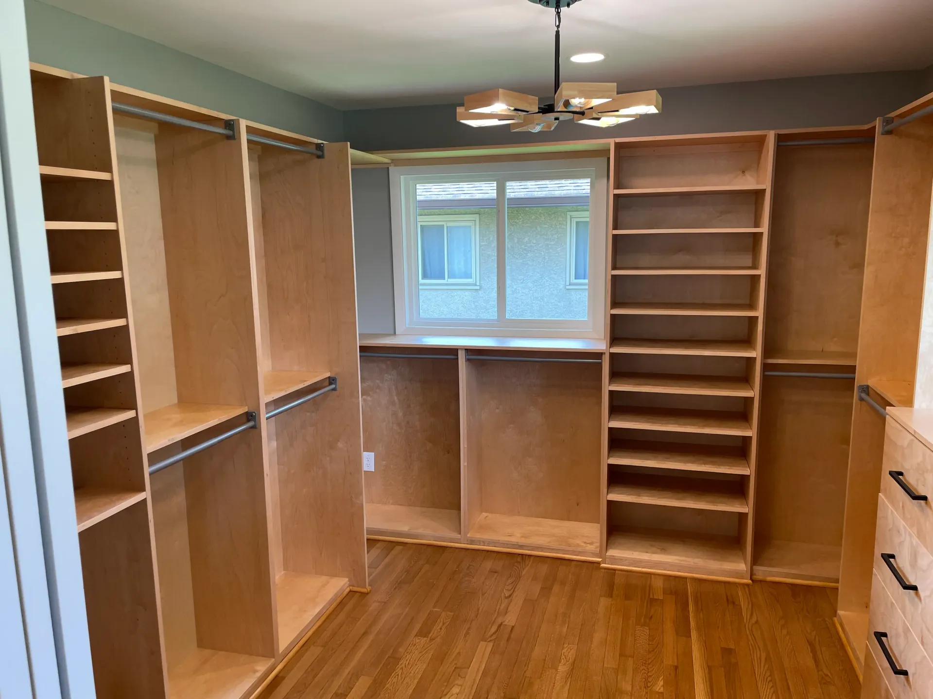 Walk-in closet with custom light wood shelving and hanging rods. Window in the center. Hardwood floor, blue-gray walls.