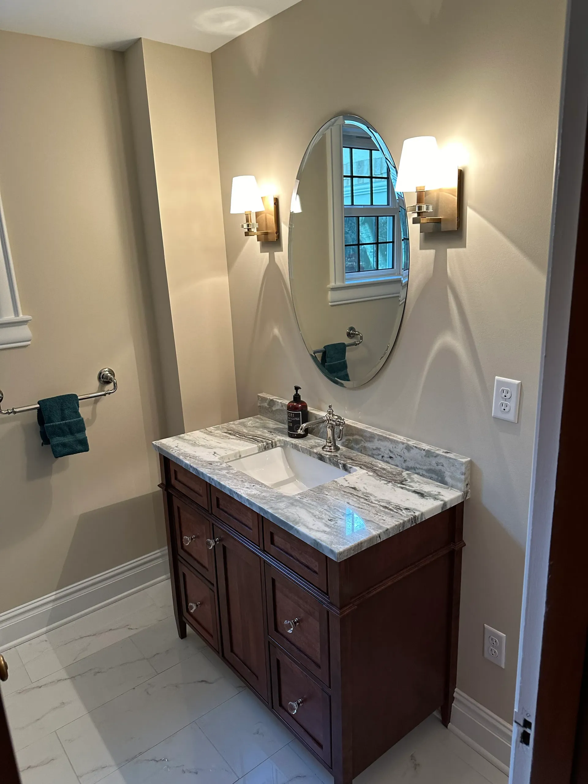 Bathroom with brown vanity, marble countertop, oval mirror, and sconce lights.