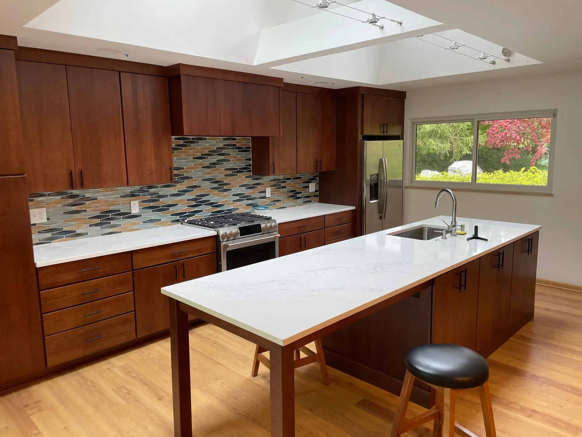 Kitchen with brown cabinets, white countertops, and a skylight. Island with sink, bar stool, and hardwood floors.