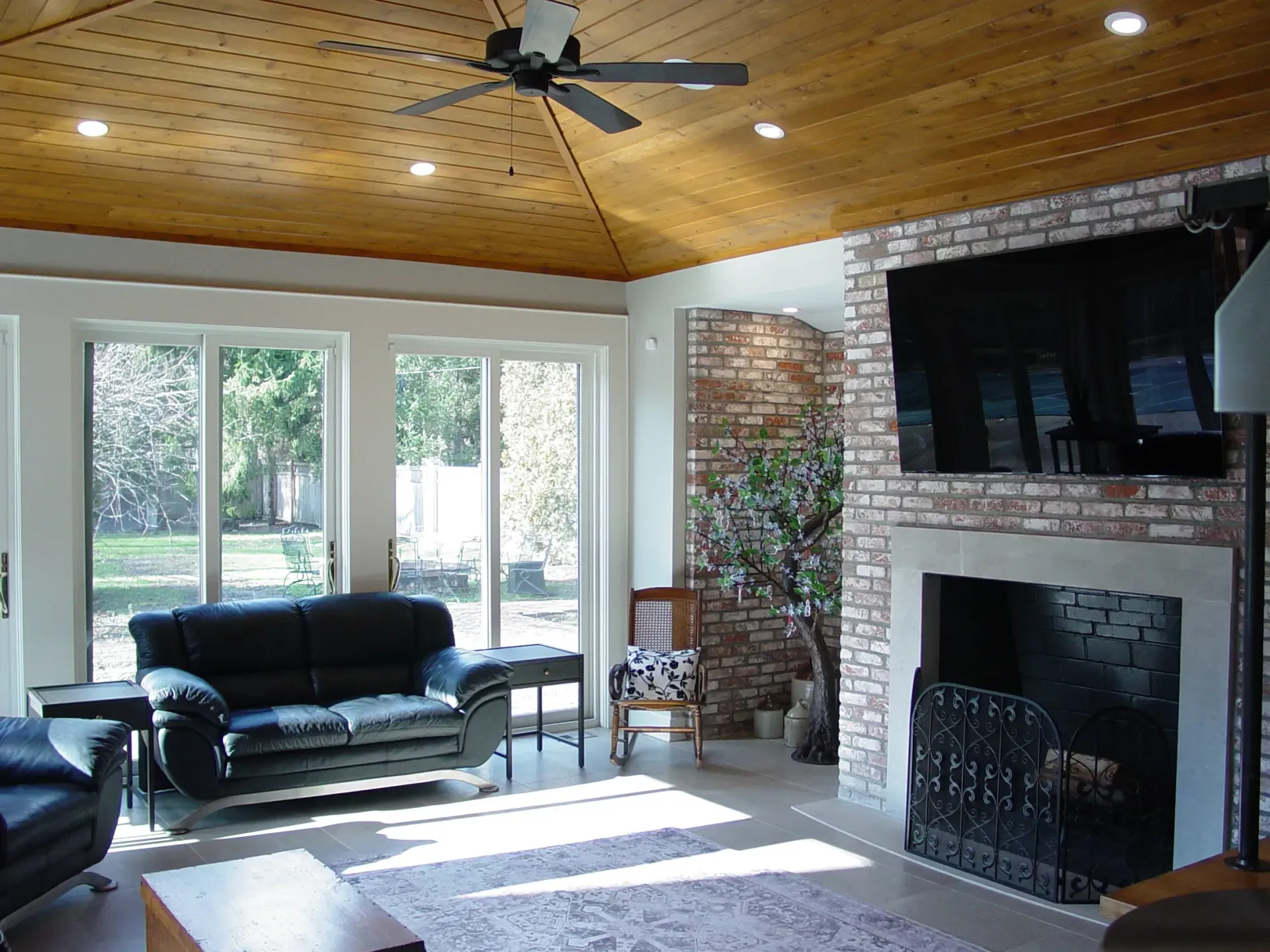 Living room with brick fireplace, black leather sofa, large windows, and wood ceiling.
