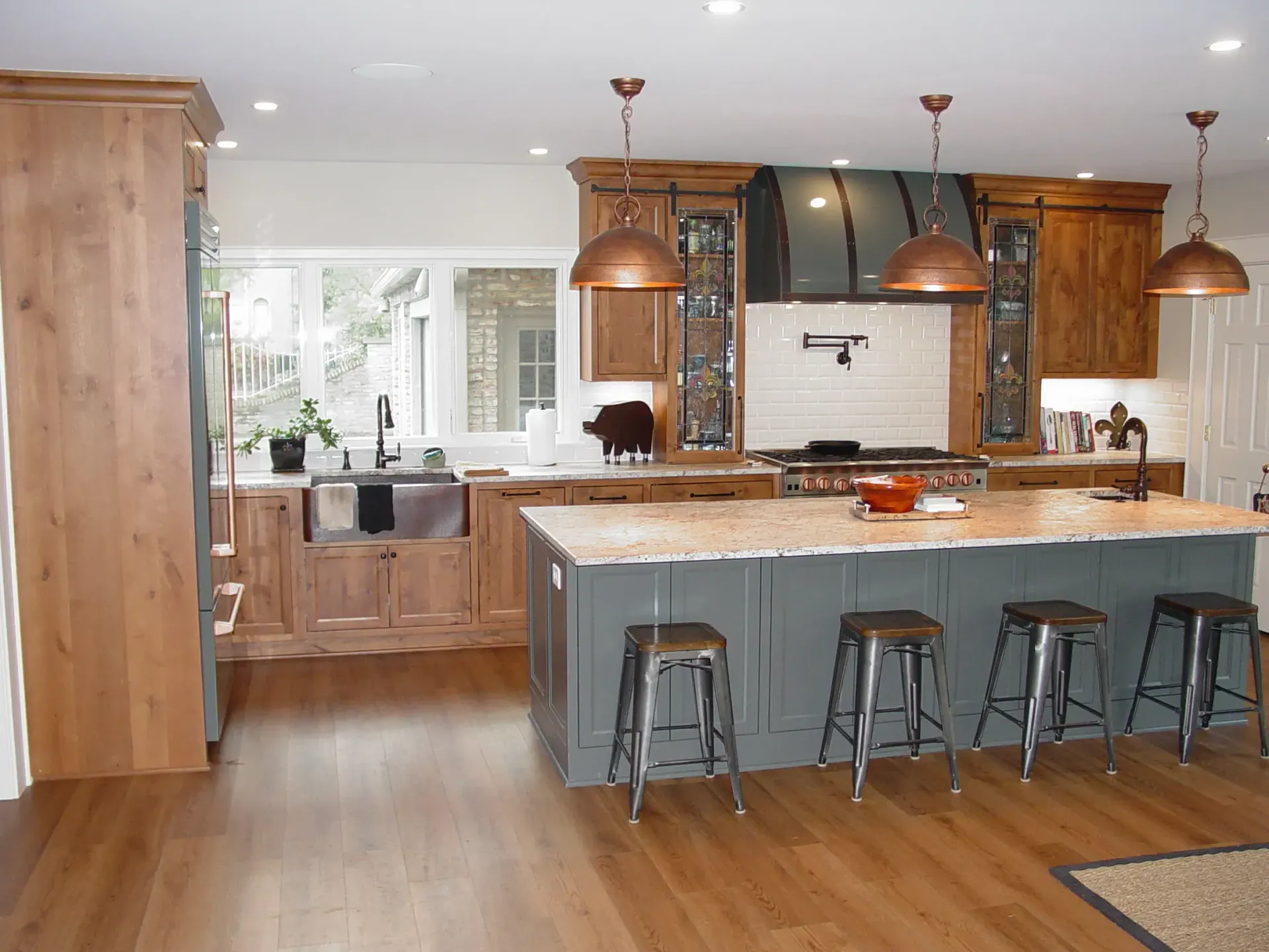 Rustic kitchen with wooden cabinets, gray island with stools, copper pendant lights, and hardwood floors.