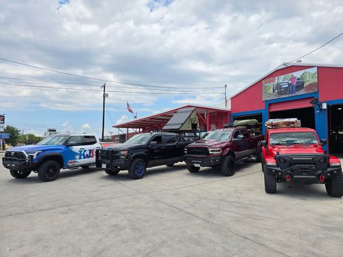 Several lifted trucks parked outside a colorful off-road shop under a cloudy sky