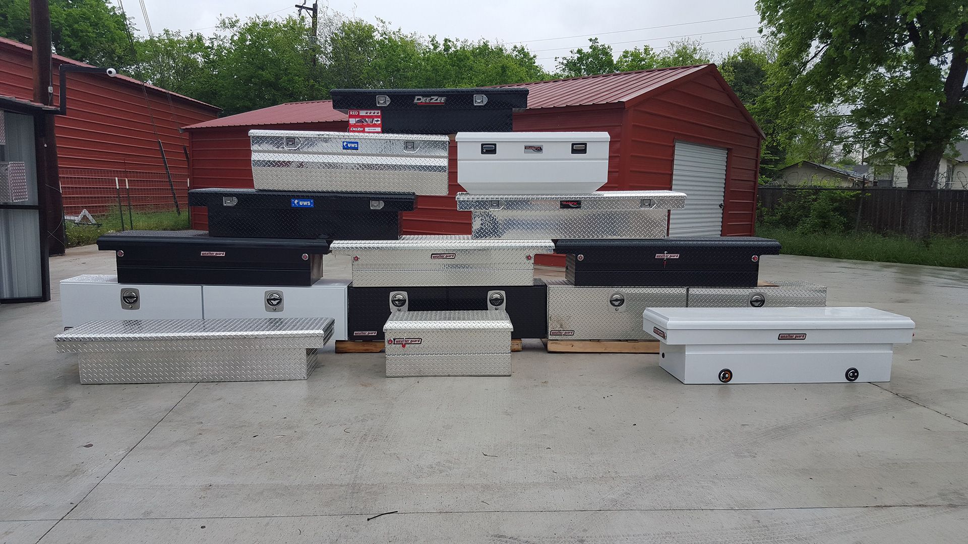Row of assorted coffins on display in an outdoor lot, with red sheds and trees in the background.
