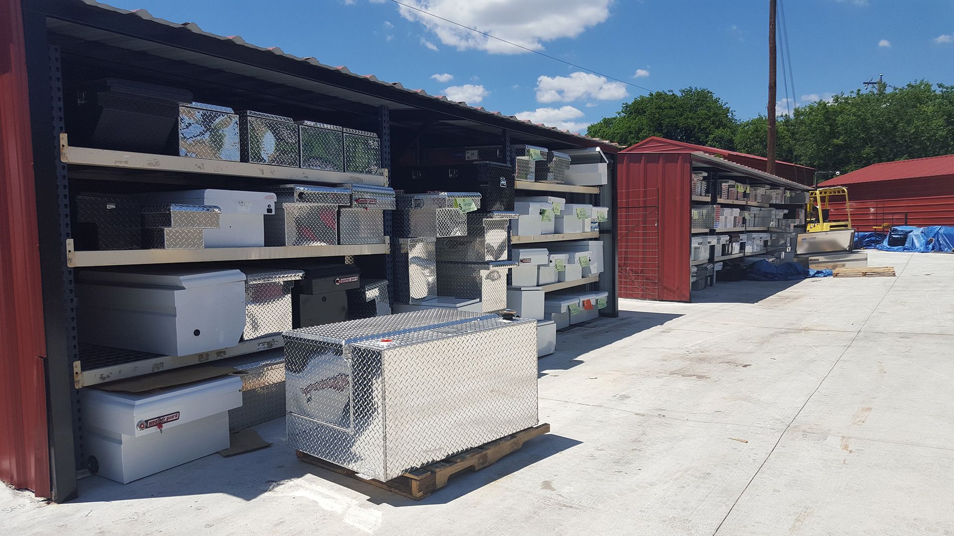 Outdoor storage area with metal shelves, stacked boxes, and a red shipping container in bright sunlight