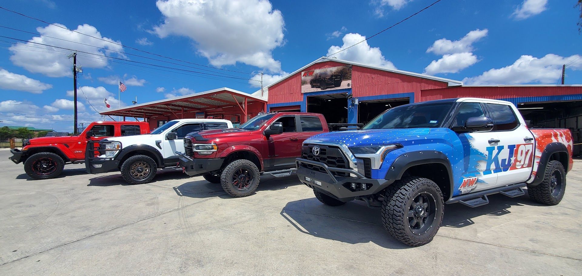 Line of off-road trucks parked outside a red building under a blue sky, including a blue race truck in front
