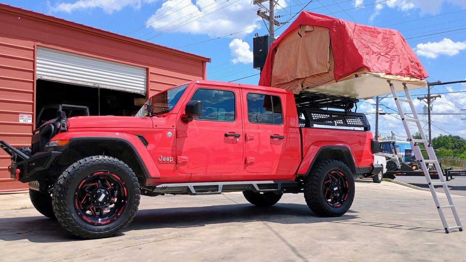 Red lifted truck with rooftop camper tent parked outside a garage on a sunny day