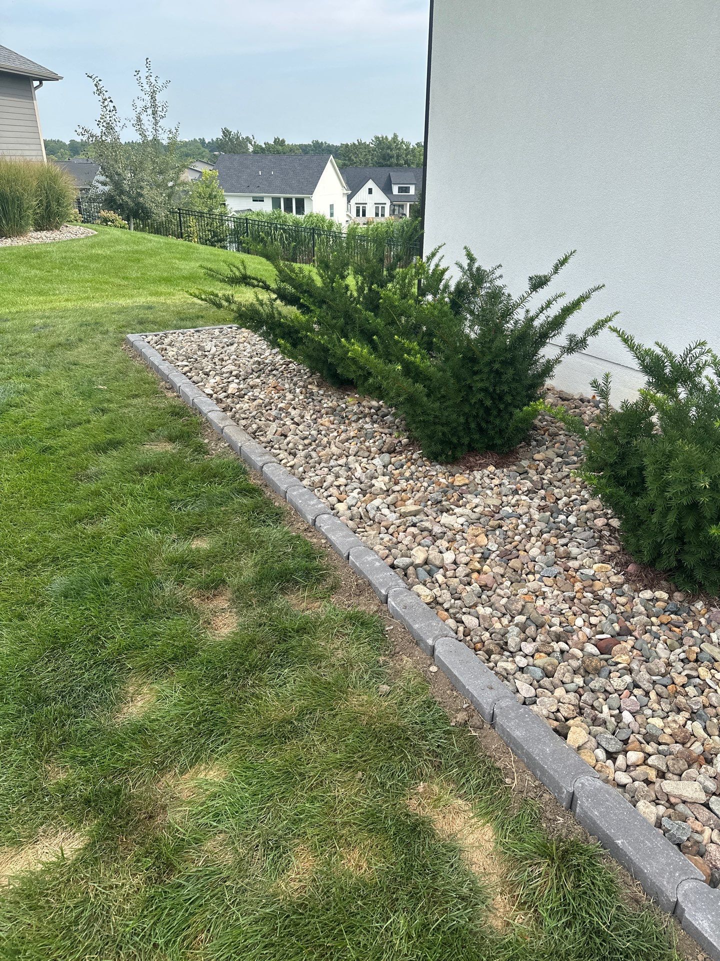 A rock garden bed with green bushes, bordered by gray pavers, next to a white wall and green lawn.