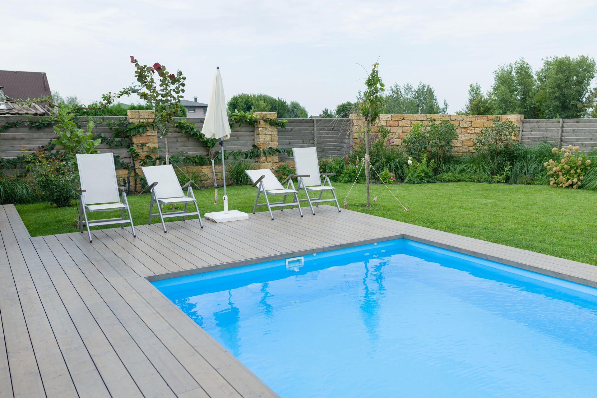 Pool with blue water, surrounded by gray deck, lawn, lounge chairs, and an umbrella on a cloudy day.