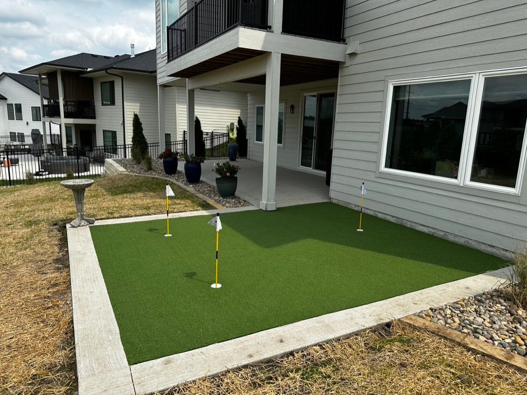 A backyard putting green with three holes and flags, surrounded by landscaping, a house, and a deck.