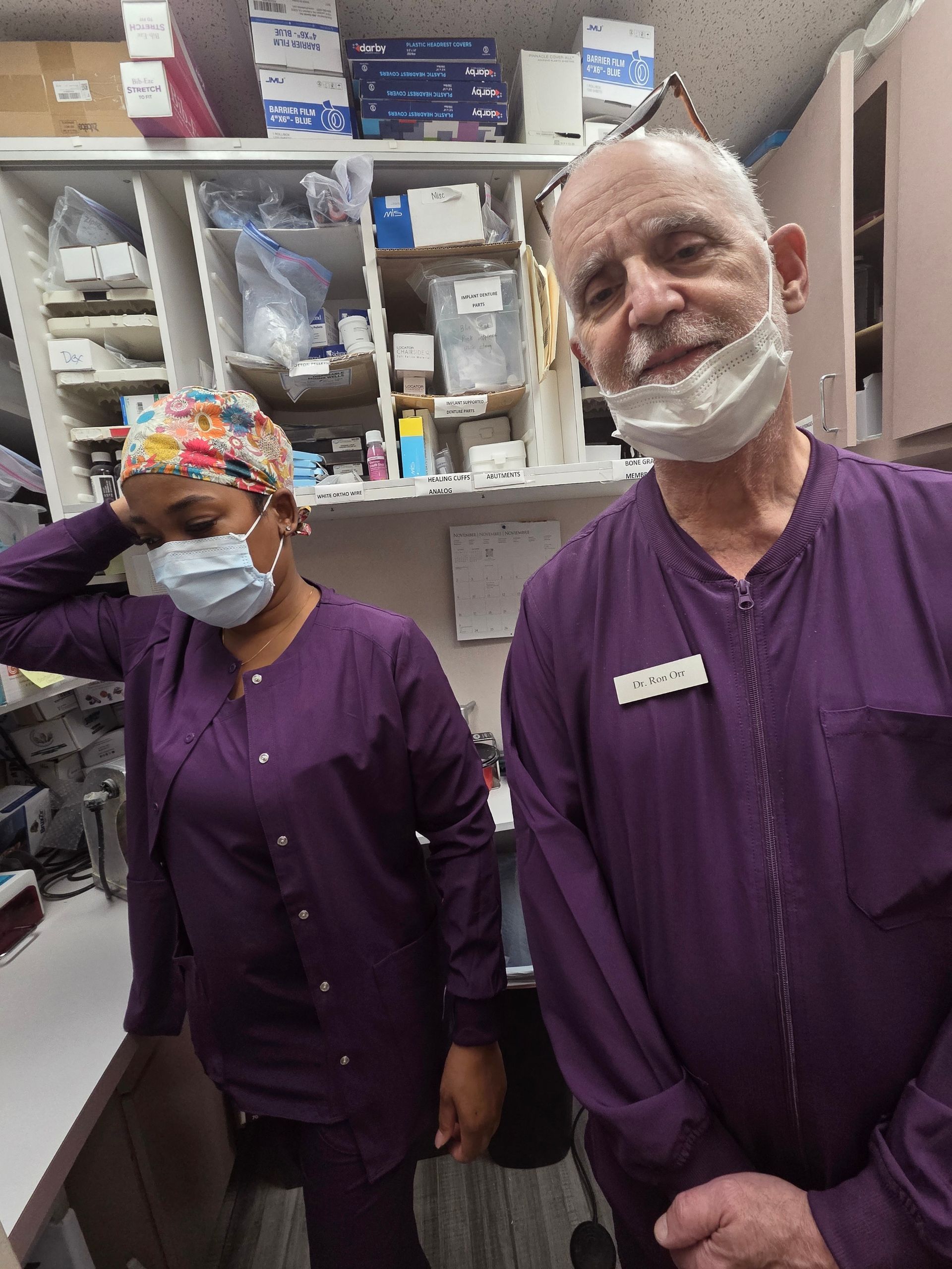 Two people in purple scrubs and masks in a medical setting, standing near shelves with supplies.