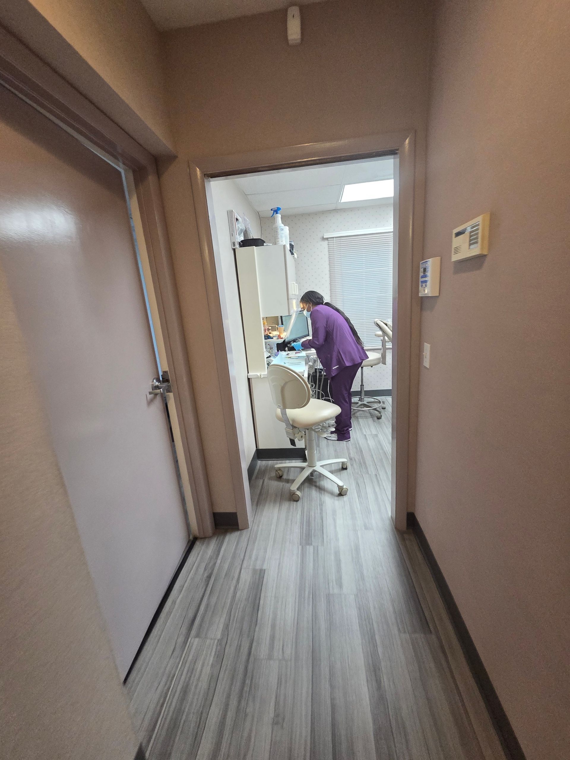Hallway with door on left, leading to dental office where a person in purple scrubs is working.