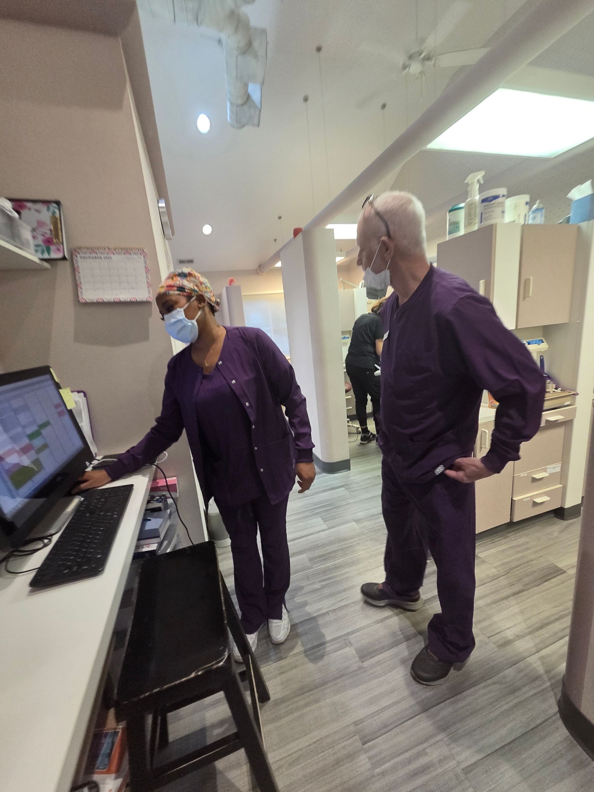 Two people in purple scrubs looking at a computer in a hallway, one wearing a mask.