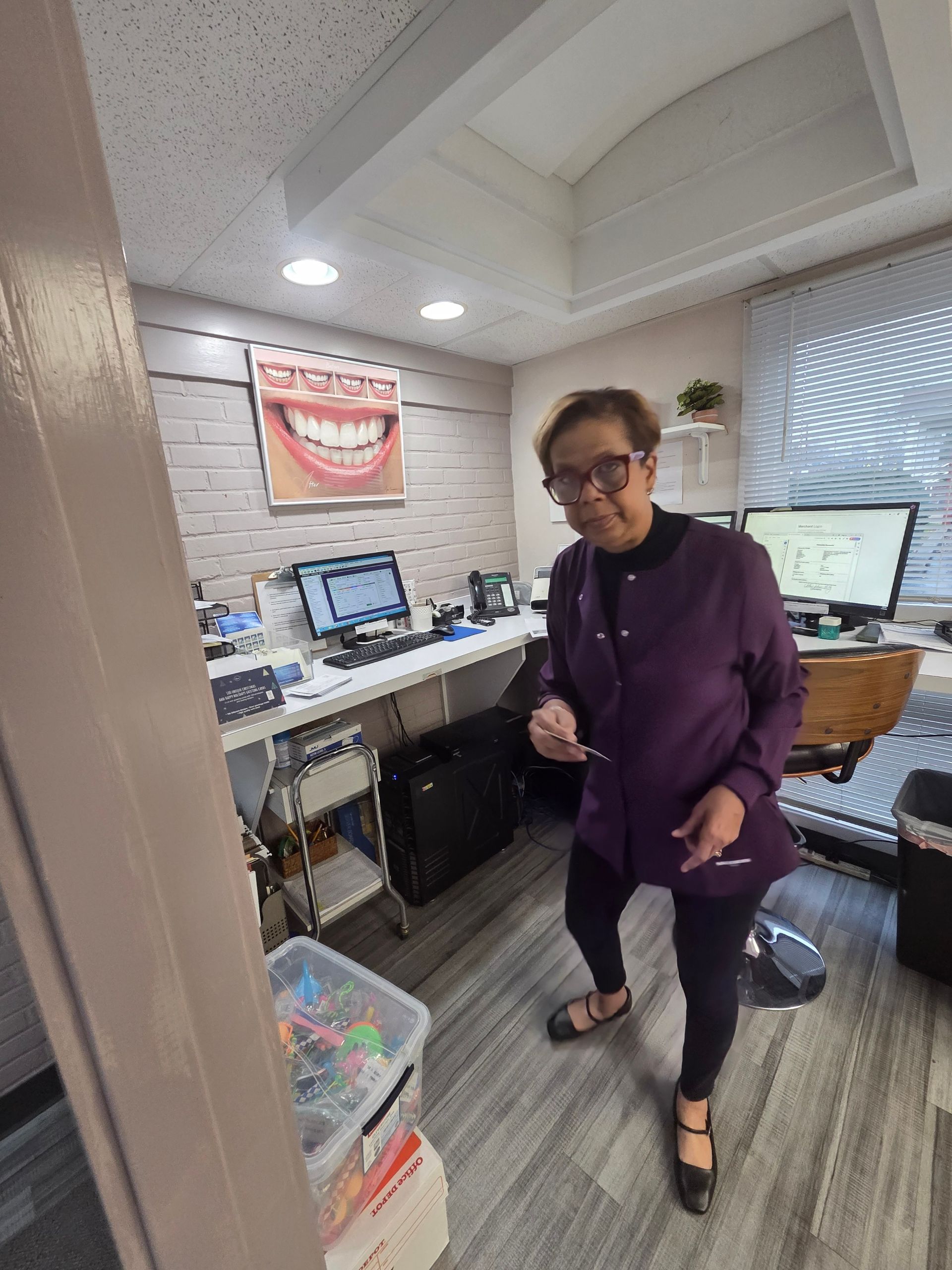 Person in purple scrubs stands in dental office, pointing. Computers, supplies, and smile diagram visible.