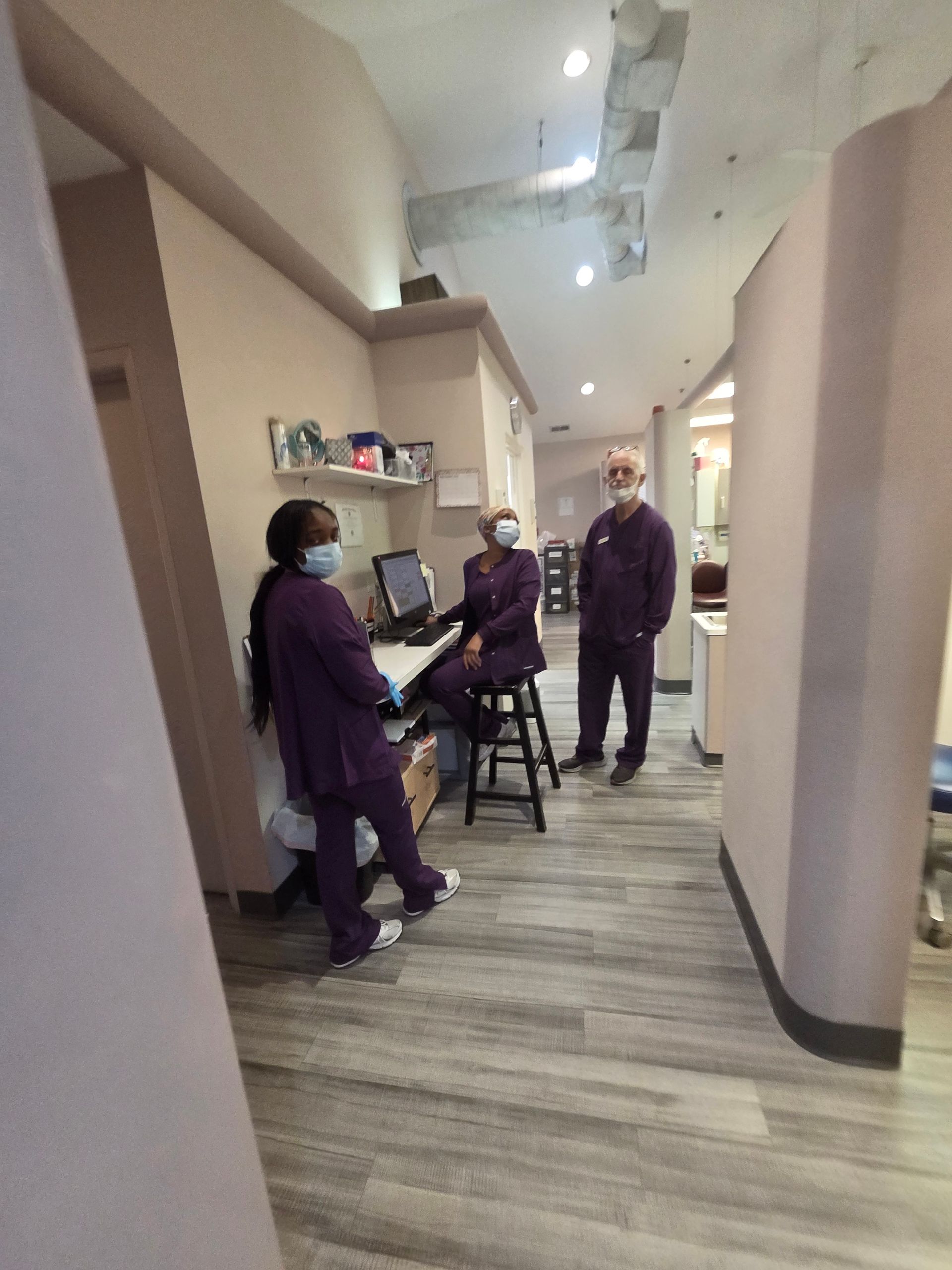 Three medical staff in a dental office hallway, wearing purple scrubs and masks.