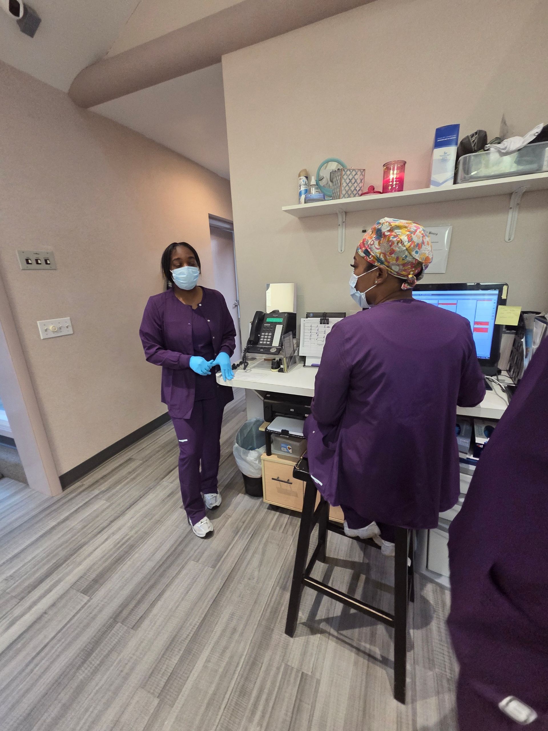 Two people in purple scrubs and masks in a medical office; one walking, one at a desk.