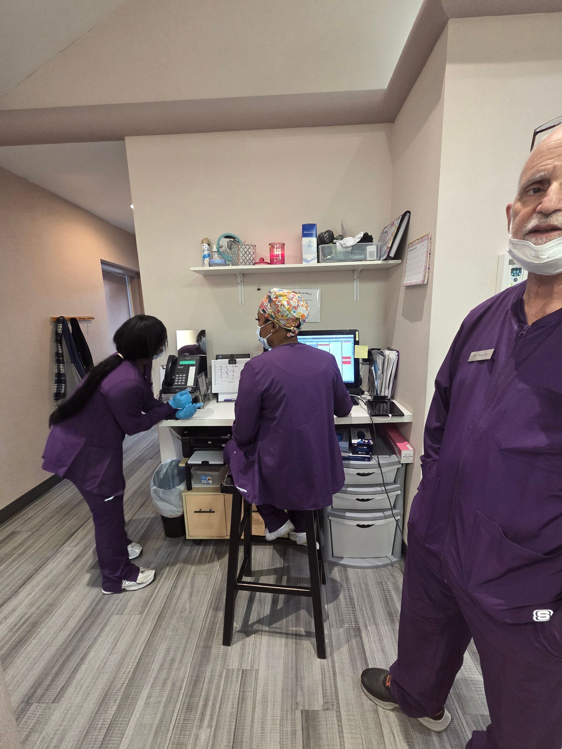 Three people in scrubs at a desk: one at a computer, one looking on, and one looking towards the camera.