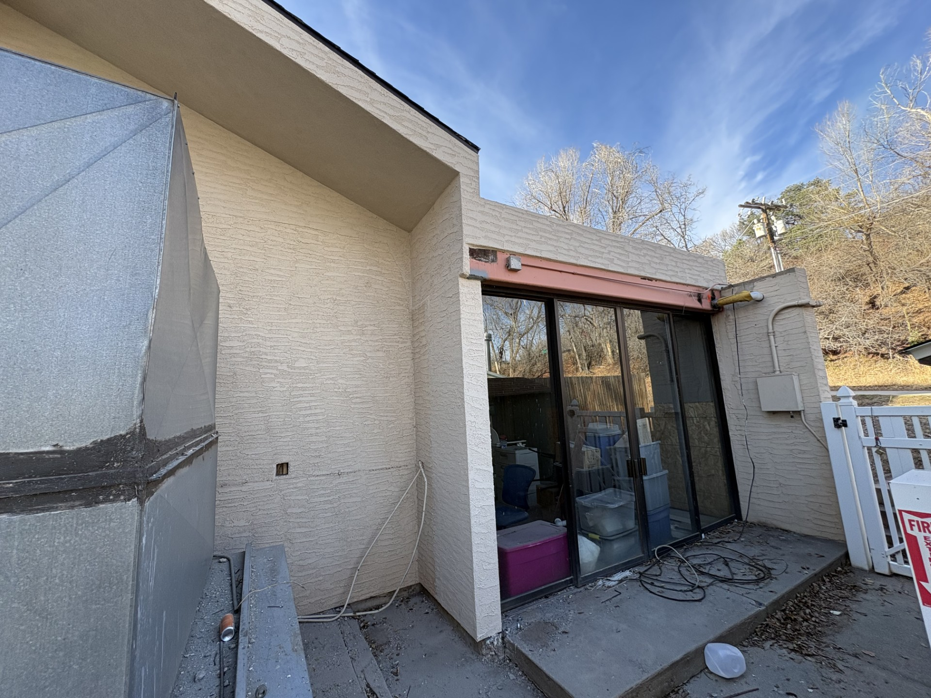 Exterior view of a building with glass sliding doors, a cement patio, and a light beige stucco exterior under a blue sky.