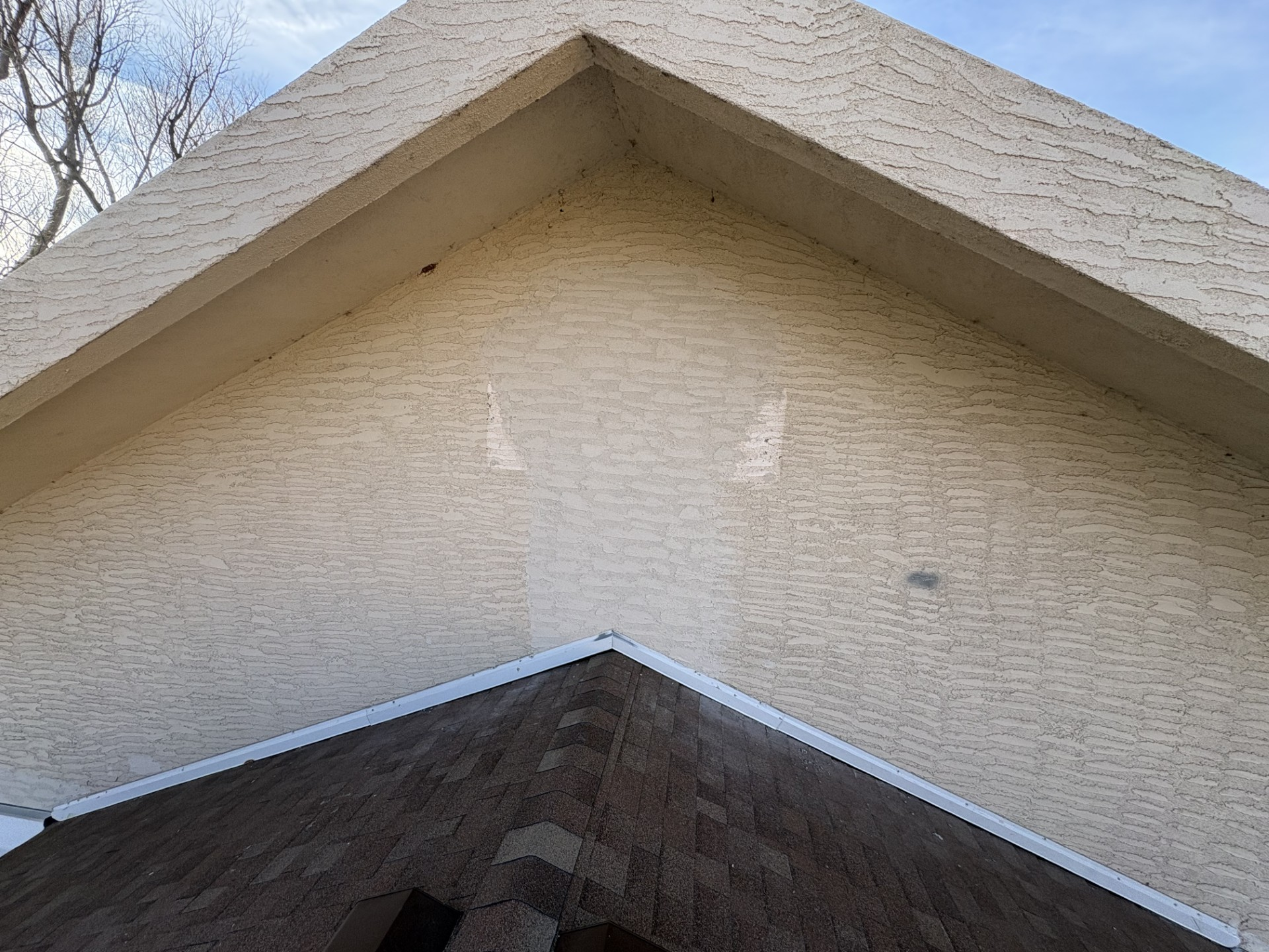 Tan stucco gable above a brown shingled roof with a blue sky background.
