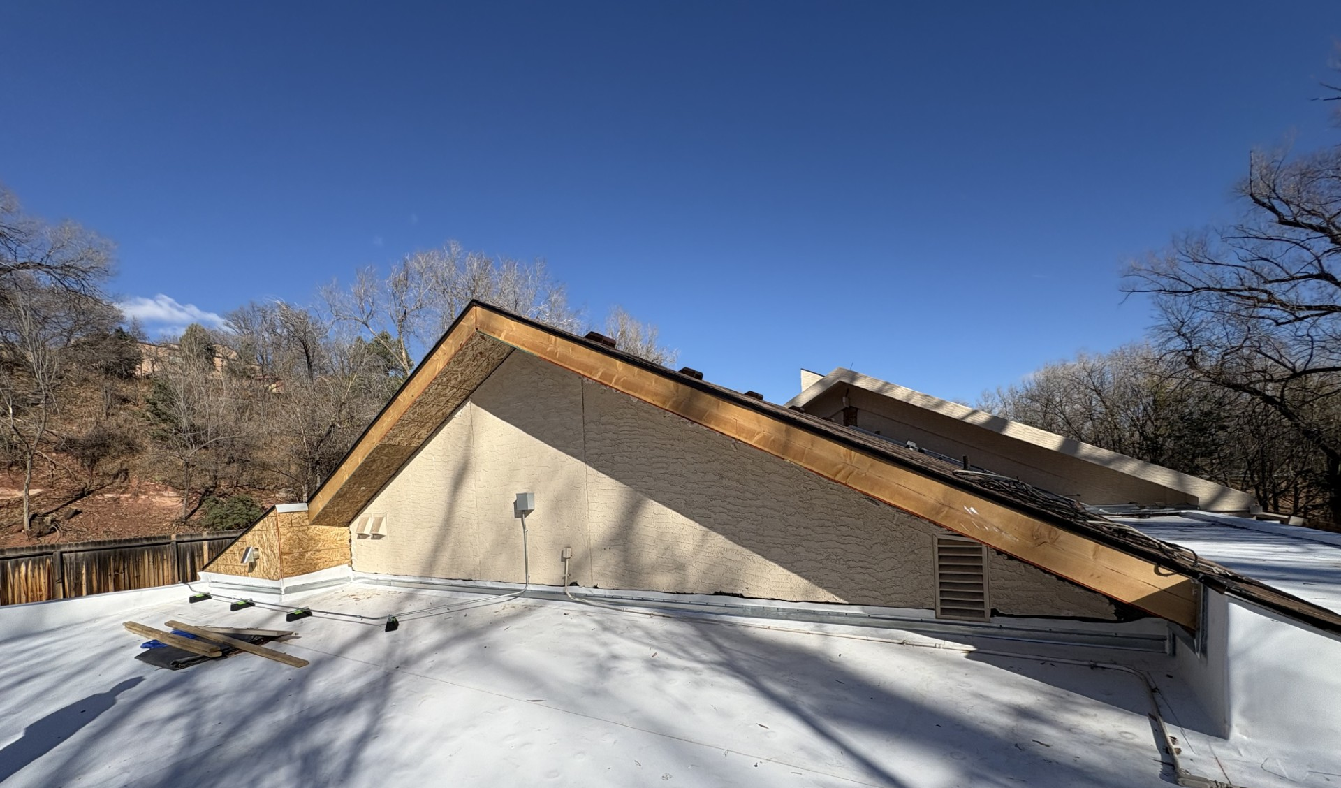 Partially snow-covered roof of a light-colored building under a clear blue sky, surrounded by bare trees.