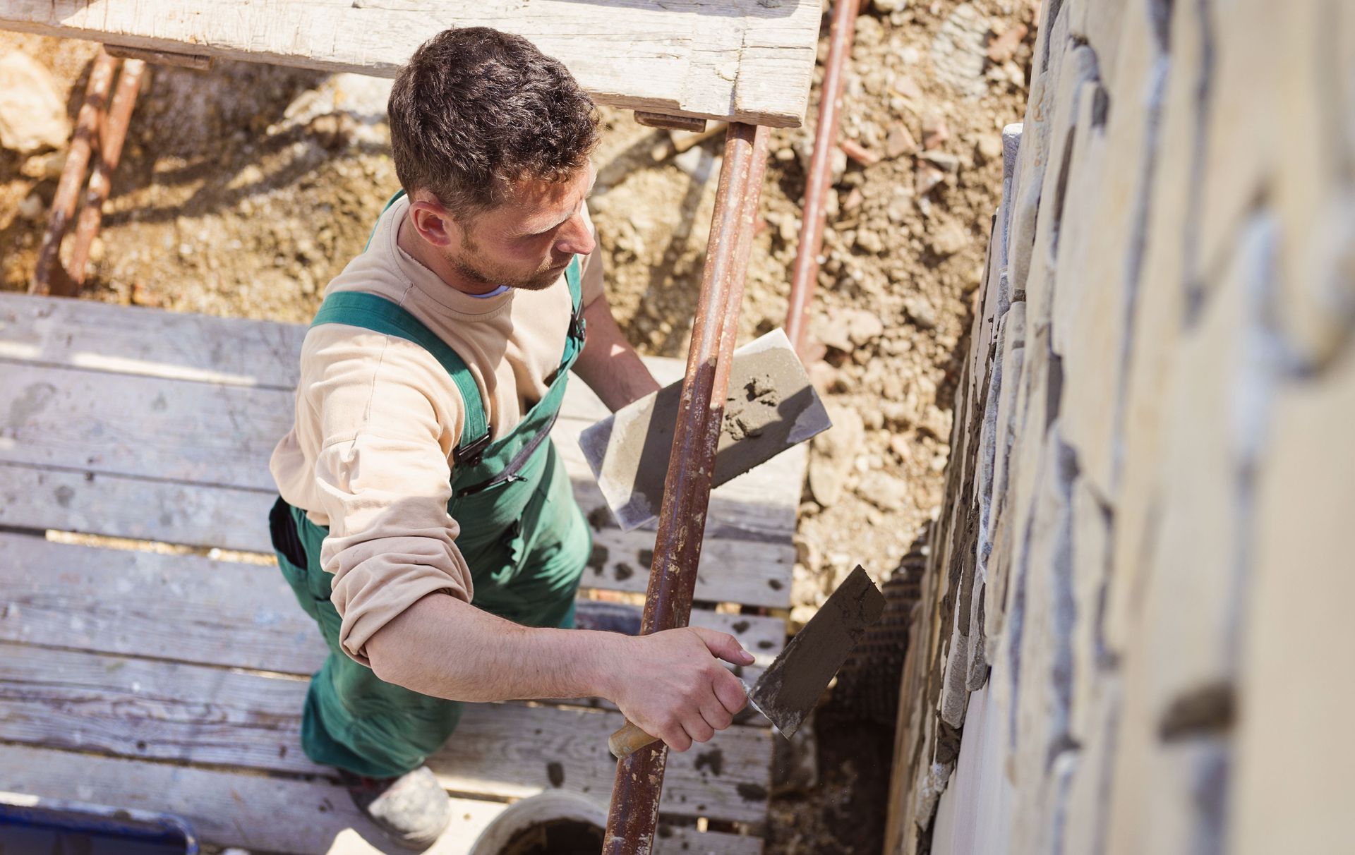 Man in green overalls applying mortar to stone wall with trowel outdoors.