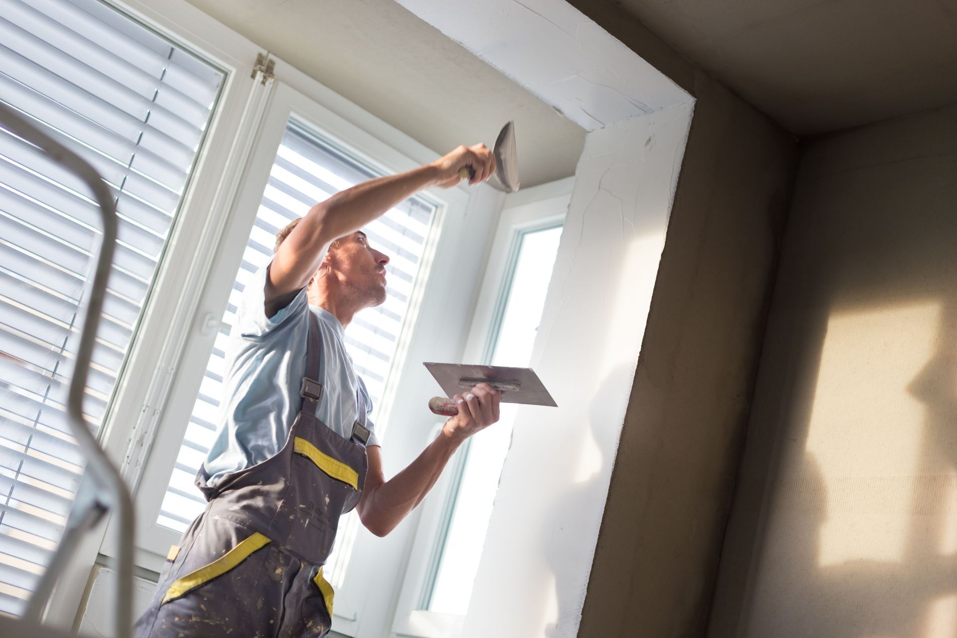 Person on ladder applying plaster to a ceiling near a window.