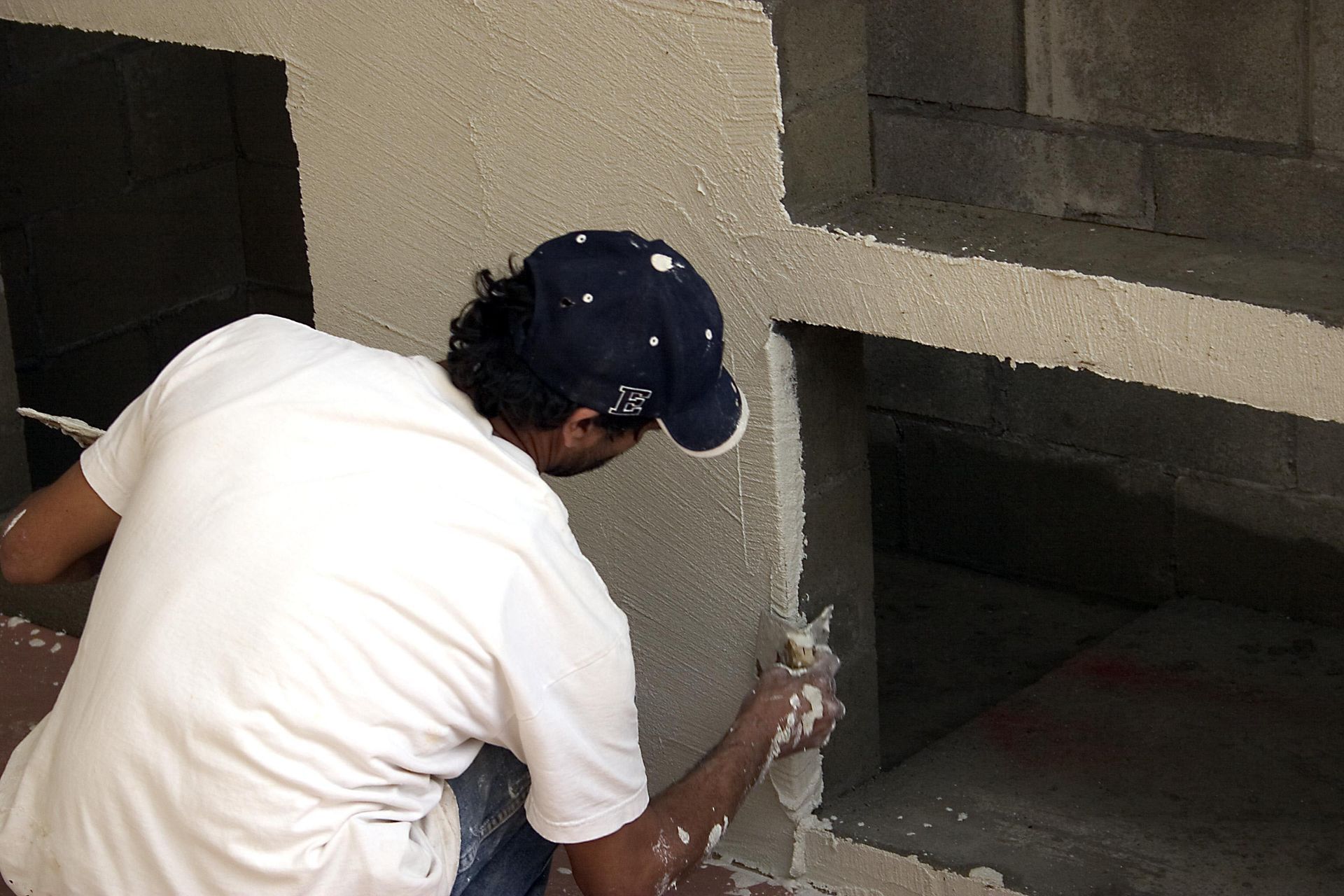 Man in white shirt and cap applying plaster to a wall with a trowel.