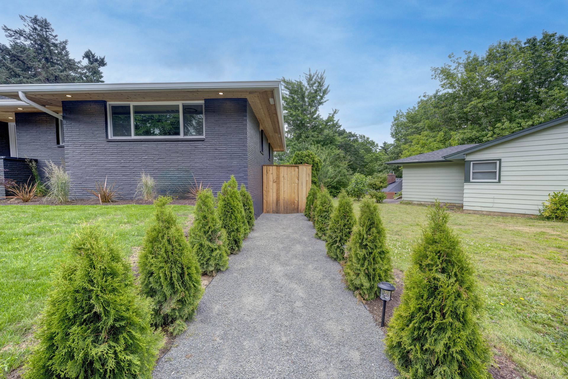 Gravel path lined with green shrubs leads to a wooden gate, between two houses on a sunny day.