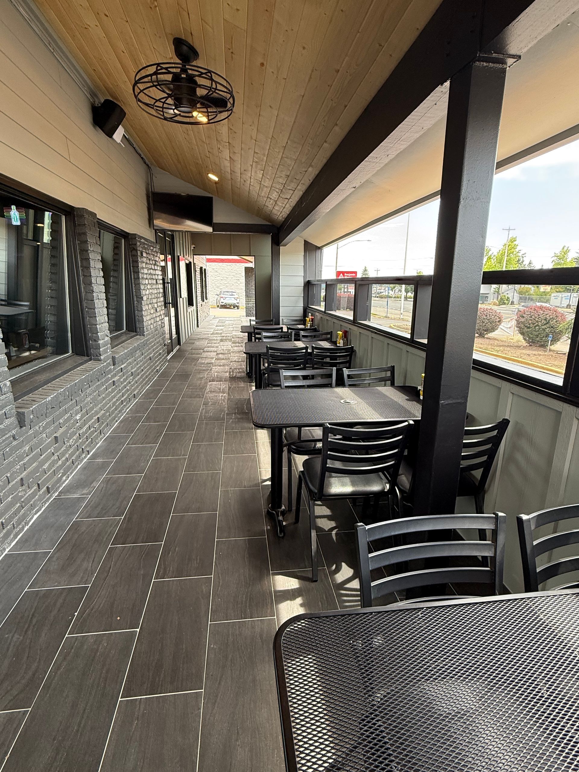 Outdoor patio with tables and chairs under a wooden ceiling; next to a wall with windows.