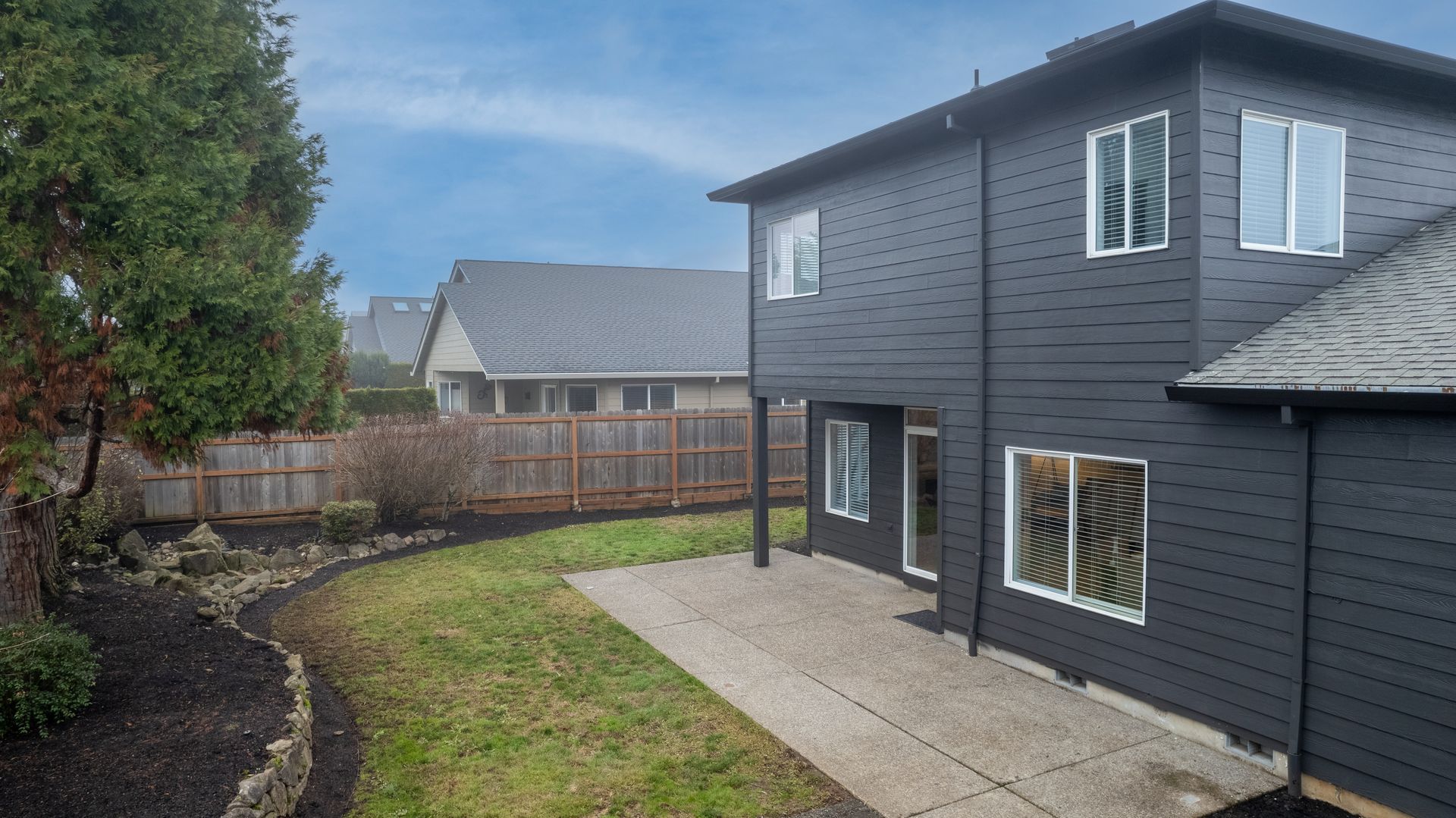 Black house with white-framed windows, gray patio, green lawn, and brown fence on a cloudy day.