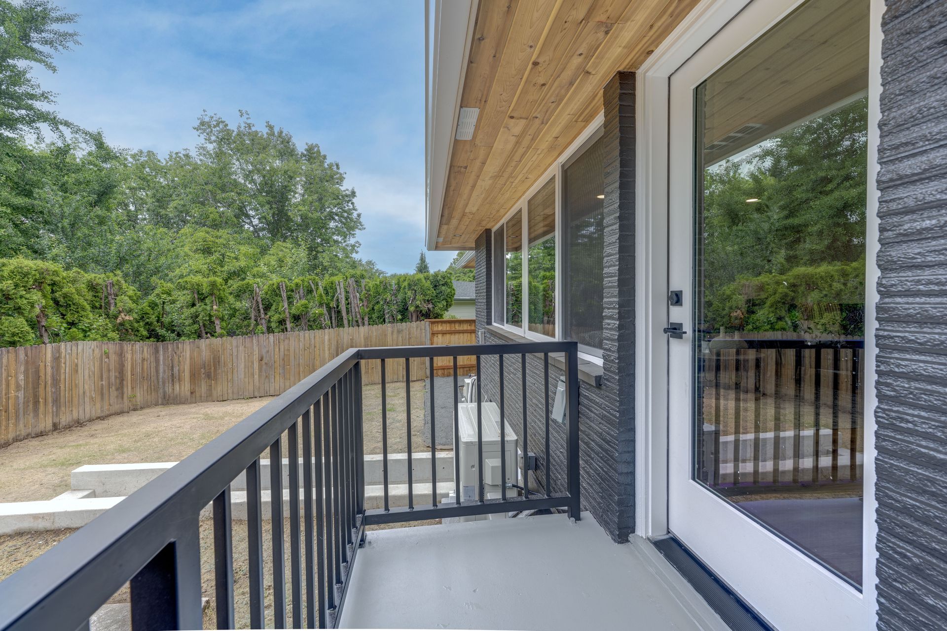 Exterior view of a modern home with black railing and textured gray walls, overlooking a yard with a fence.