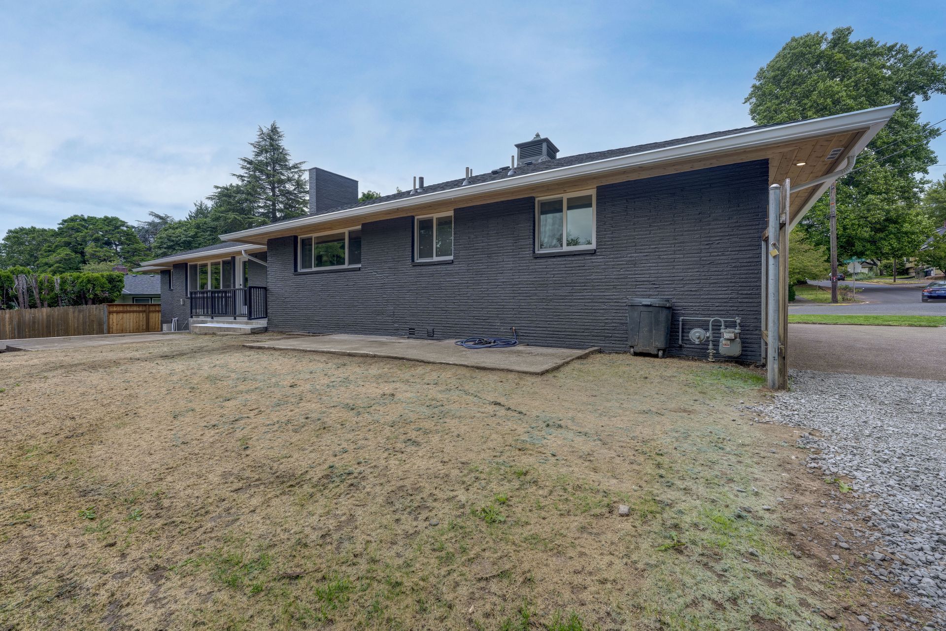 Back view of a dark-sided house with light-colored roof, standing on a grassy yard under a cloudy sky.