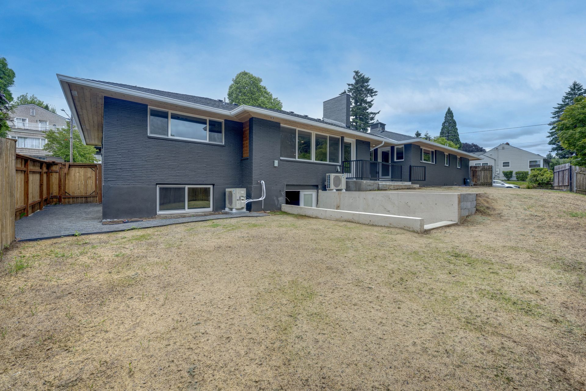 Back of a dark gray house with a dry lawn, a wooden fence, and blue sky.