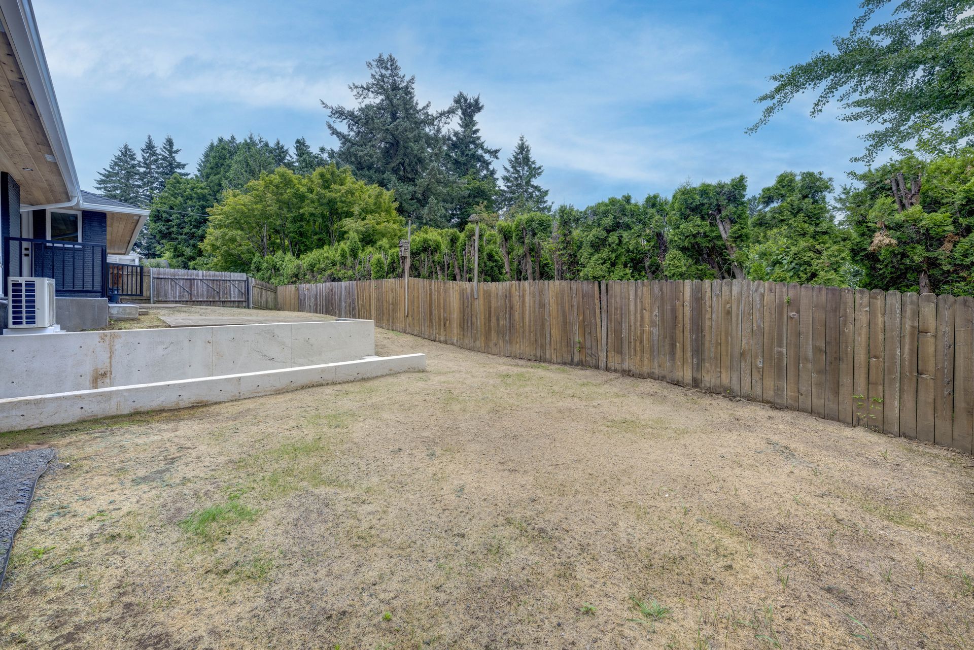 Backyard with wooden fence, dry grass, concrete wall, and trees under a partly cloudy sky.