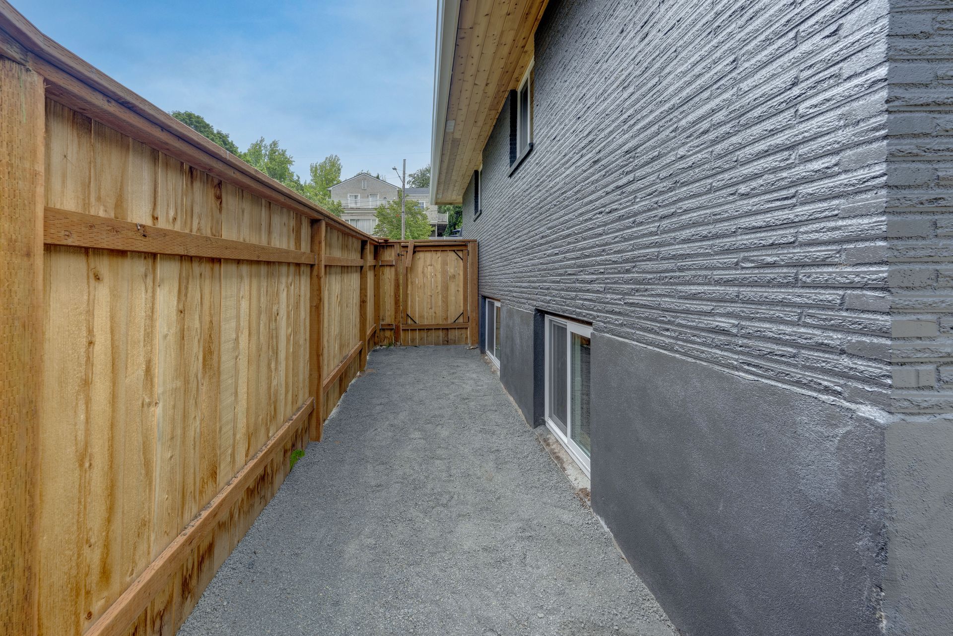 Narrow, gravel pathway between a wooden fence and dark gray building.