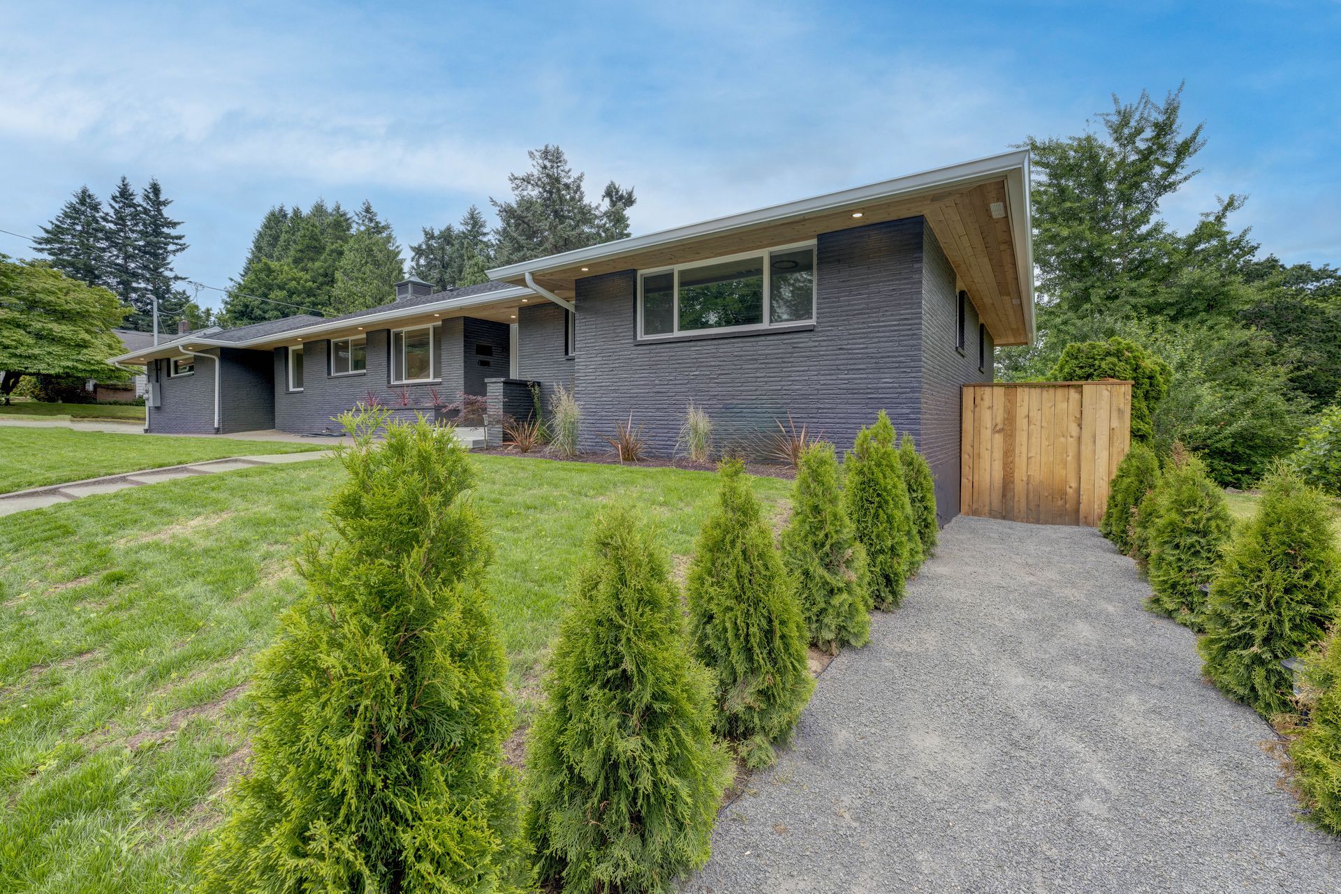 A gray brick ranch house with a gravel path lined by evergreen trees, under a blue sky.
