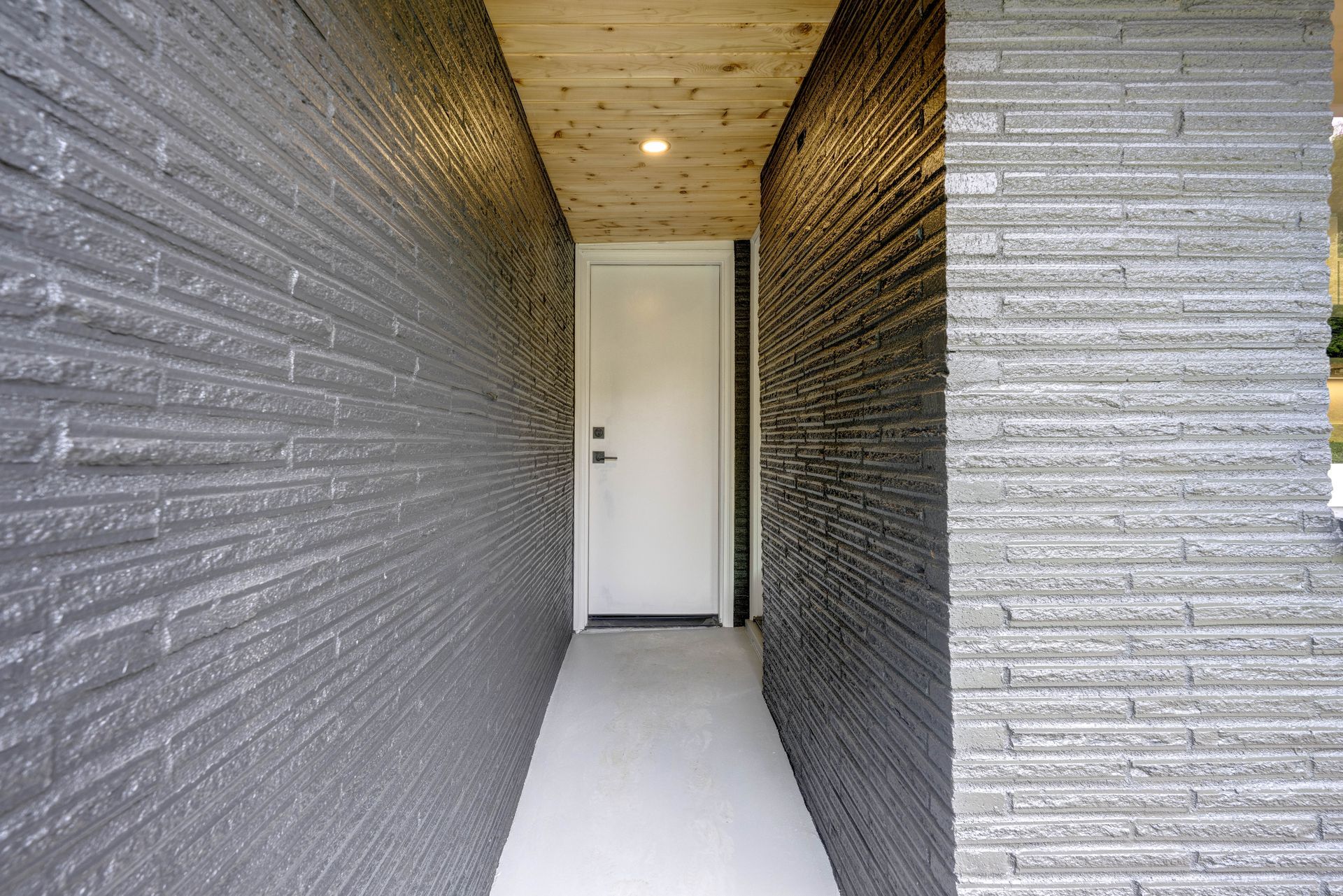 A narrow entryway with grey brick walls leading to a white door under a wooden ceiling.