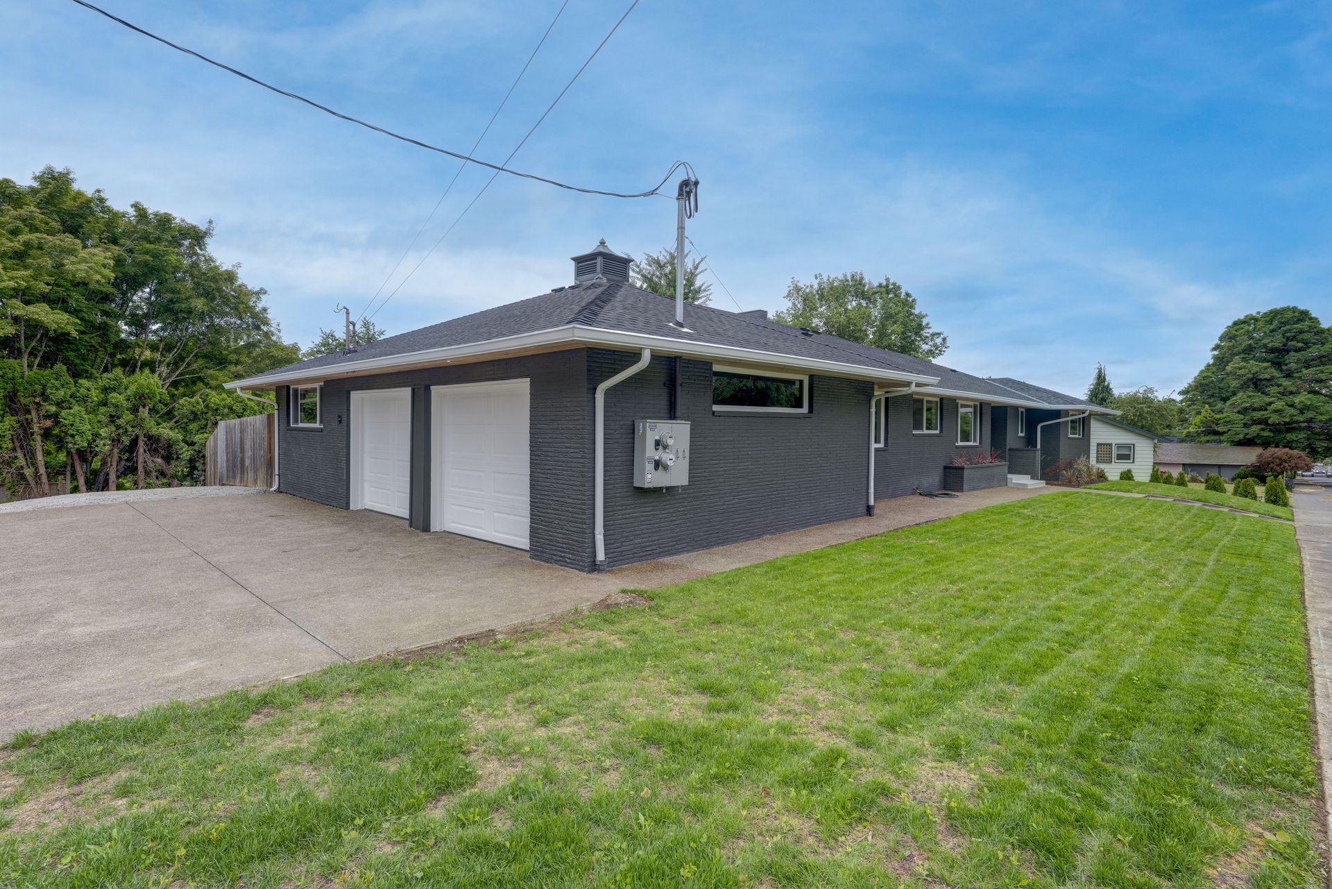 Dark gray house with two-car garage, green lawn, and concrete driveway under a blue sky.