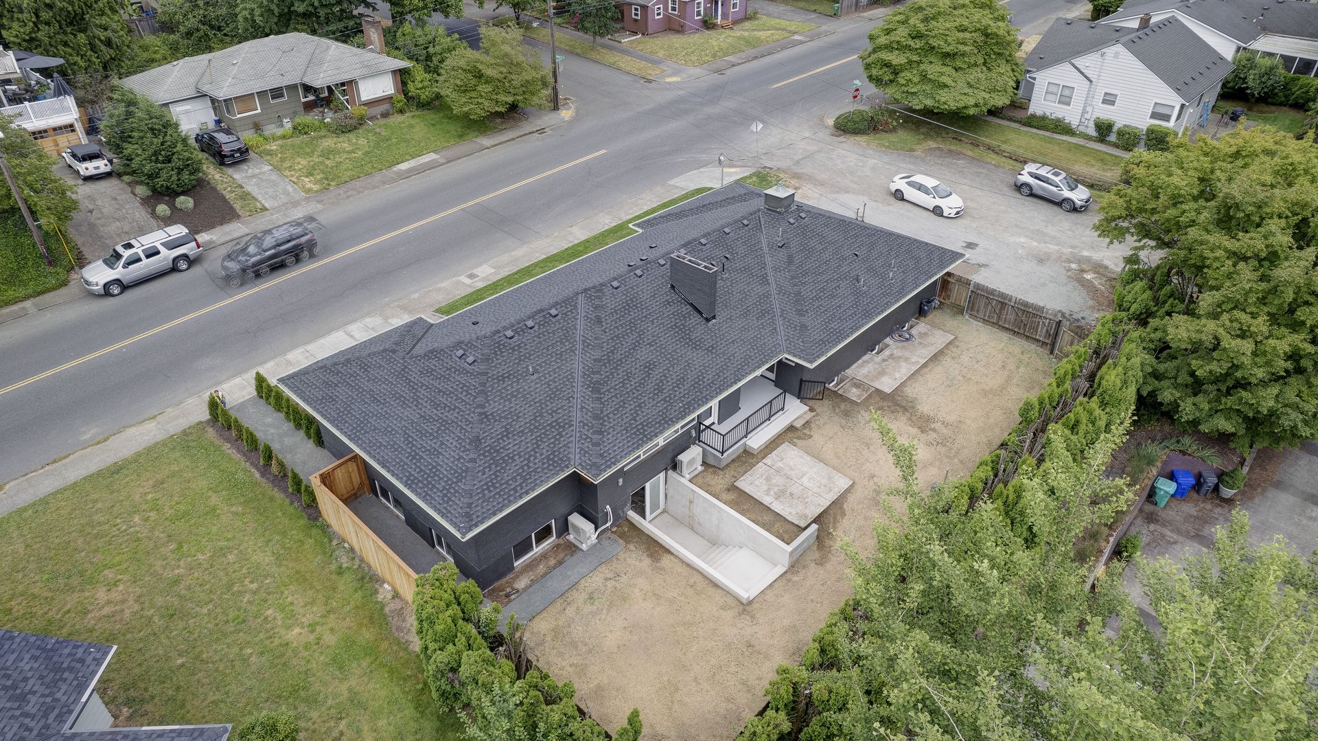 Aerial view of a dark gray house with a black roof, surrounded by a fence, near a street with cars.