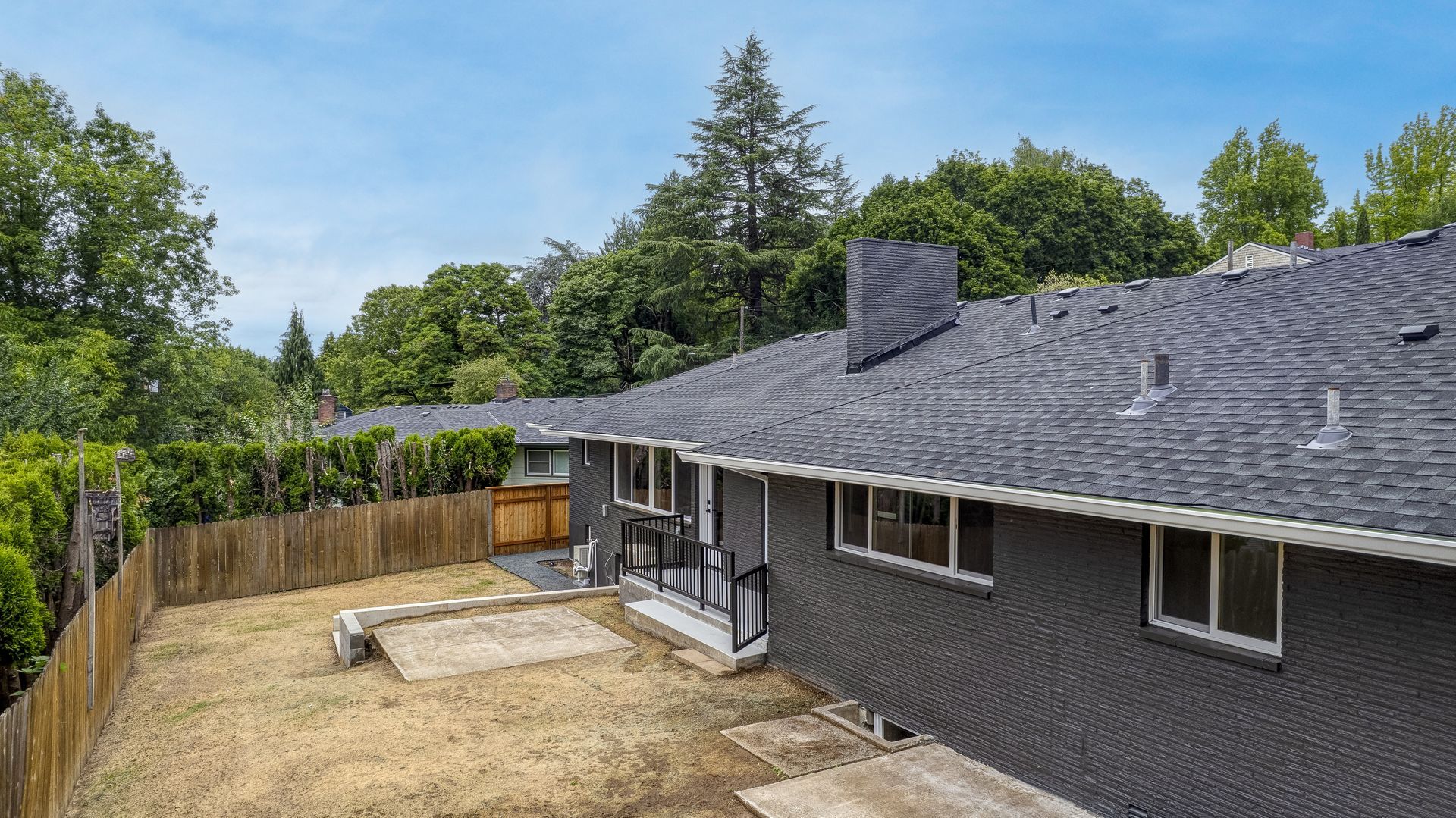 Backyard view of a dark gray house with a chimney, brown fence, and patches of dry grass.