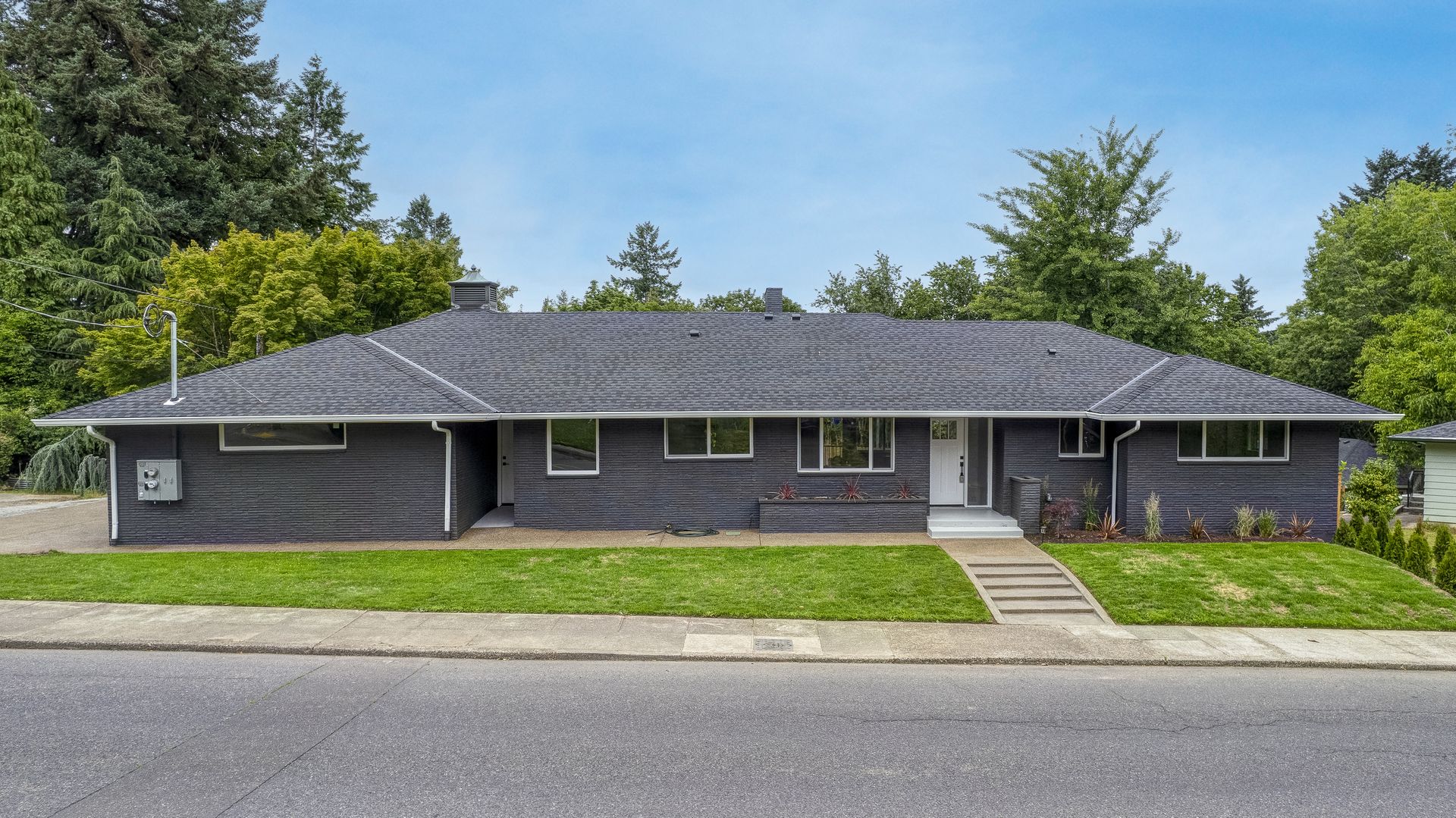 Single-story house with dark exterior and gray tiled roof, set on a lawn, sidewalk, and street.