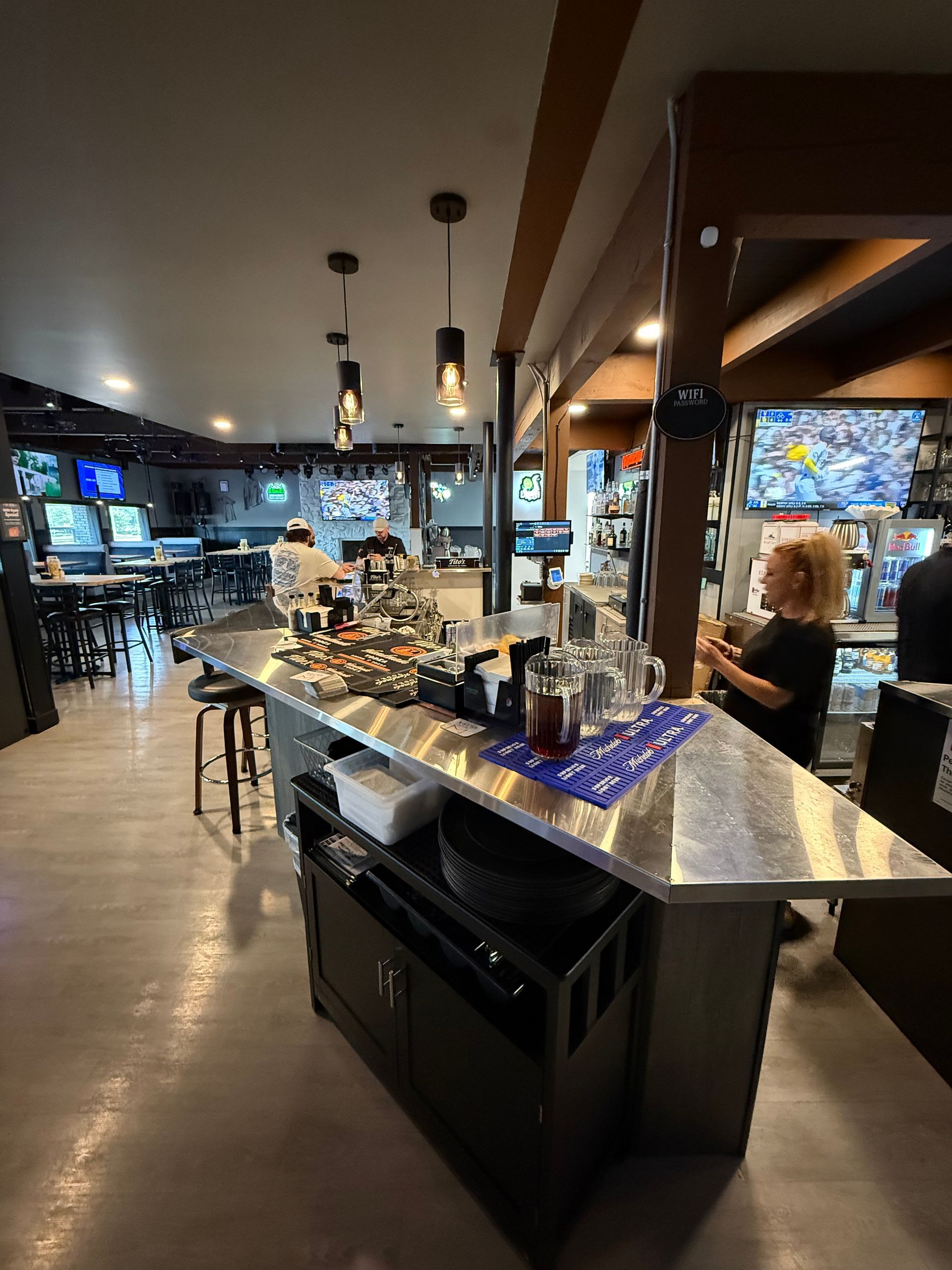A bar interior with a bartender, bar stools, drinks, and multiple TVs.