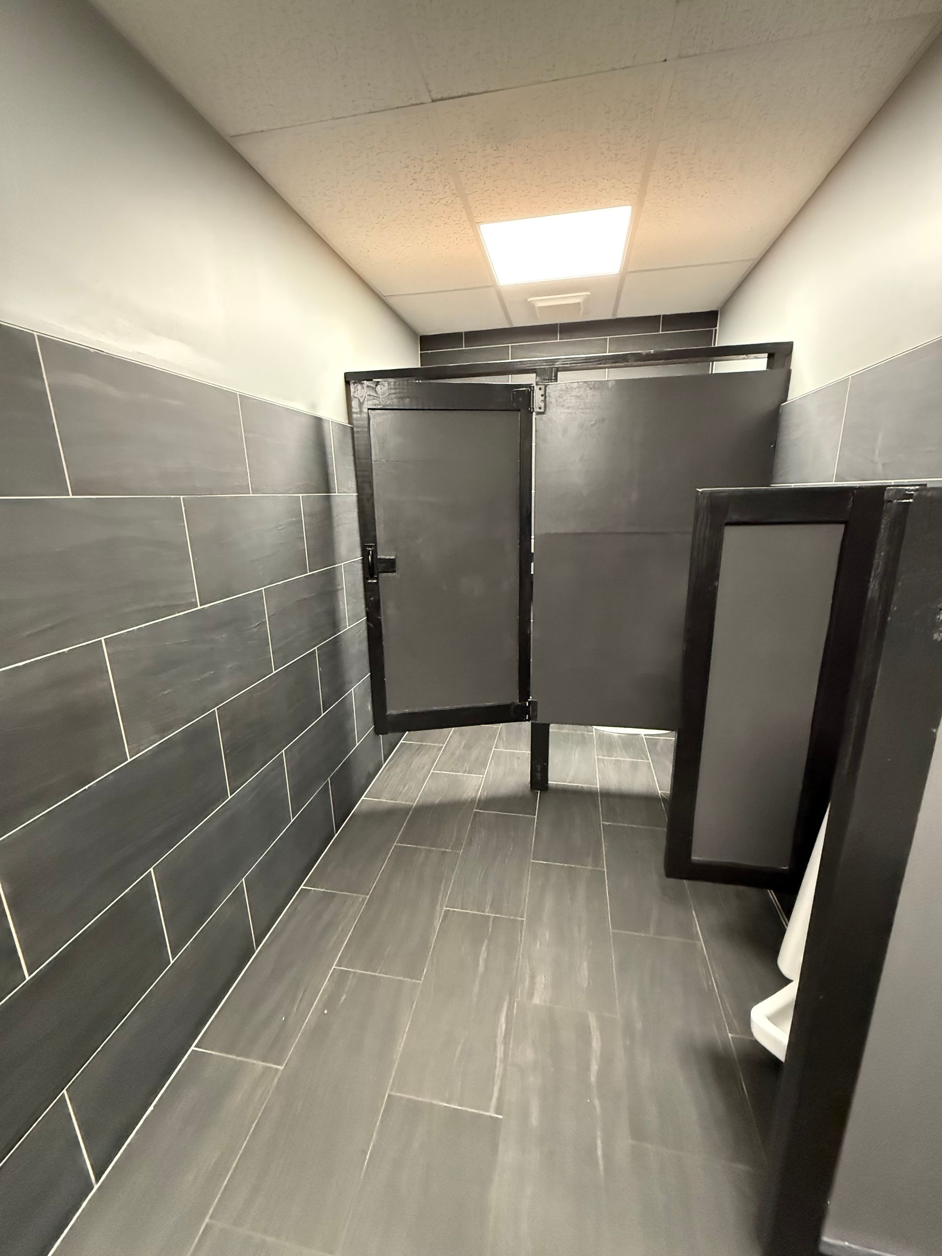 Public restroom with gray tile walls and stalls; fluorescent lighting.