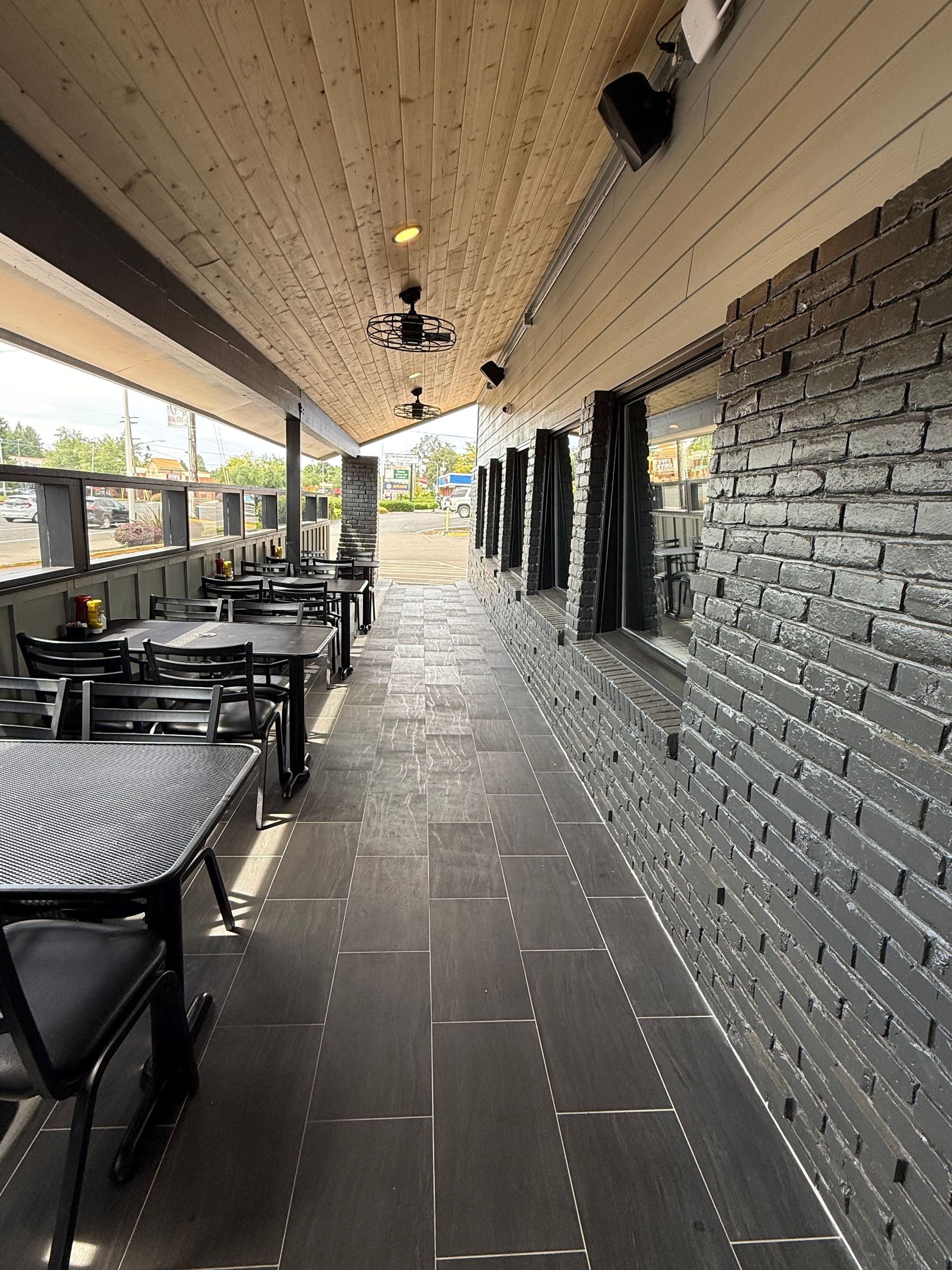 Outdoor restaurant patio with black tables and chairs, gray brick wall, wooden ceiling, and stone floor.