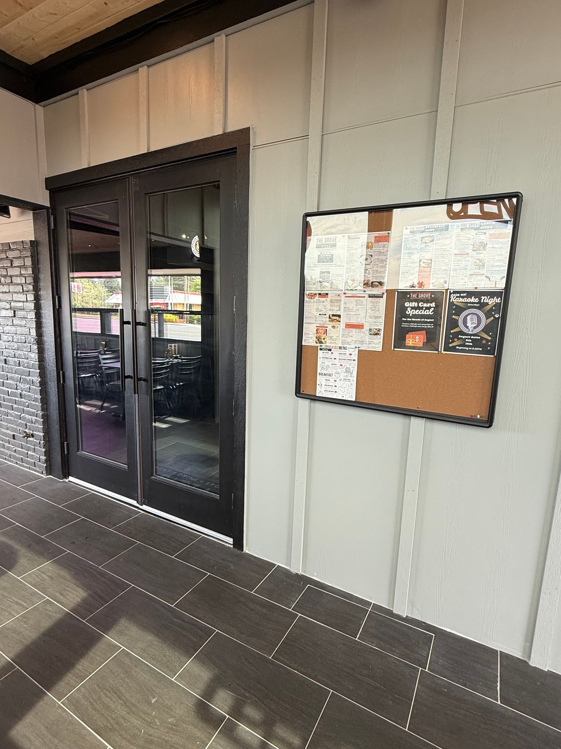 Exterior entrance with double doors, menu board on the wall. Gray building with dark wood doors and a tiled floor.